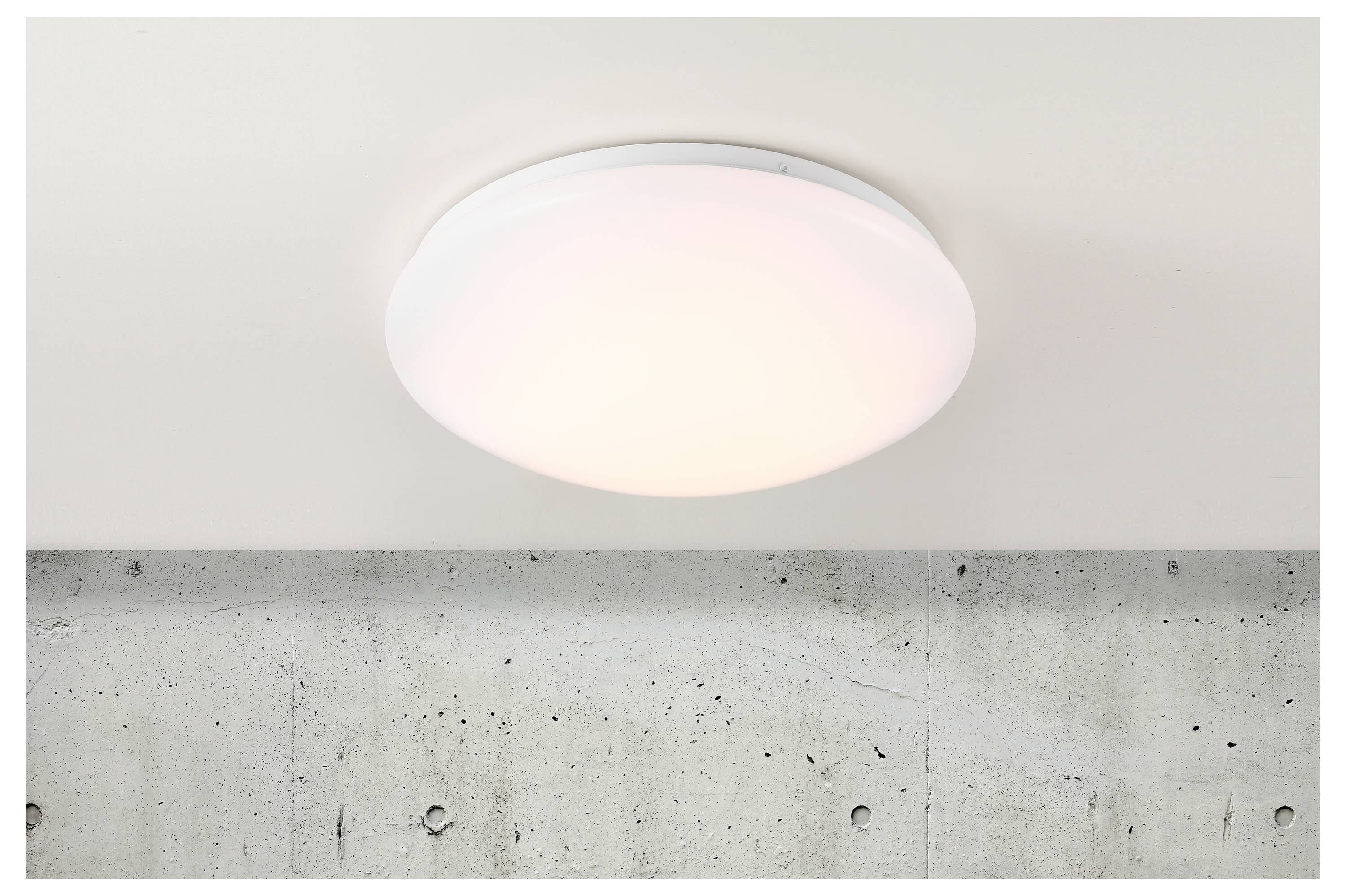 Deckenleuchte, die auf einer weißen Decke angebracht ist, über einer Betonwand. Die Leuchte gibt ein weiches, weißes Licht ab und zeigt an, dass sie eingeschaltet ist.