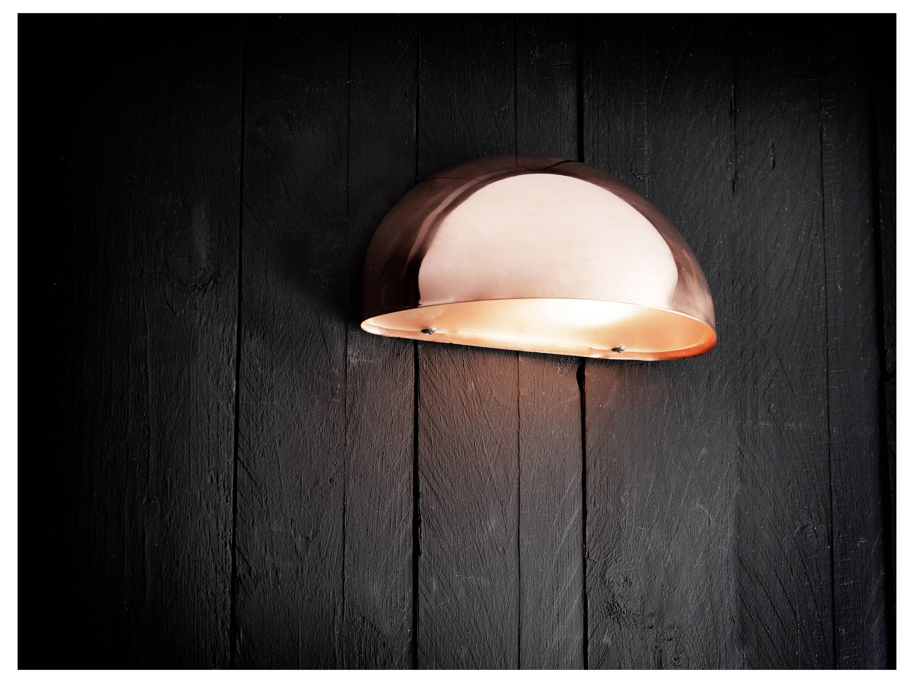 Eine kupferne Halbkuppel-Wandleuchte strahlt warmes Licht gegen eine dunkle Holzwand aus.