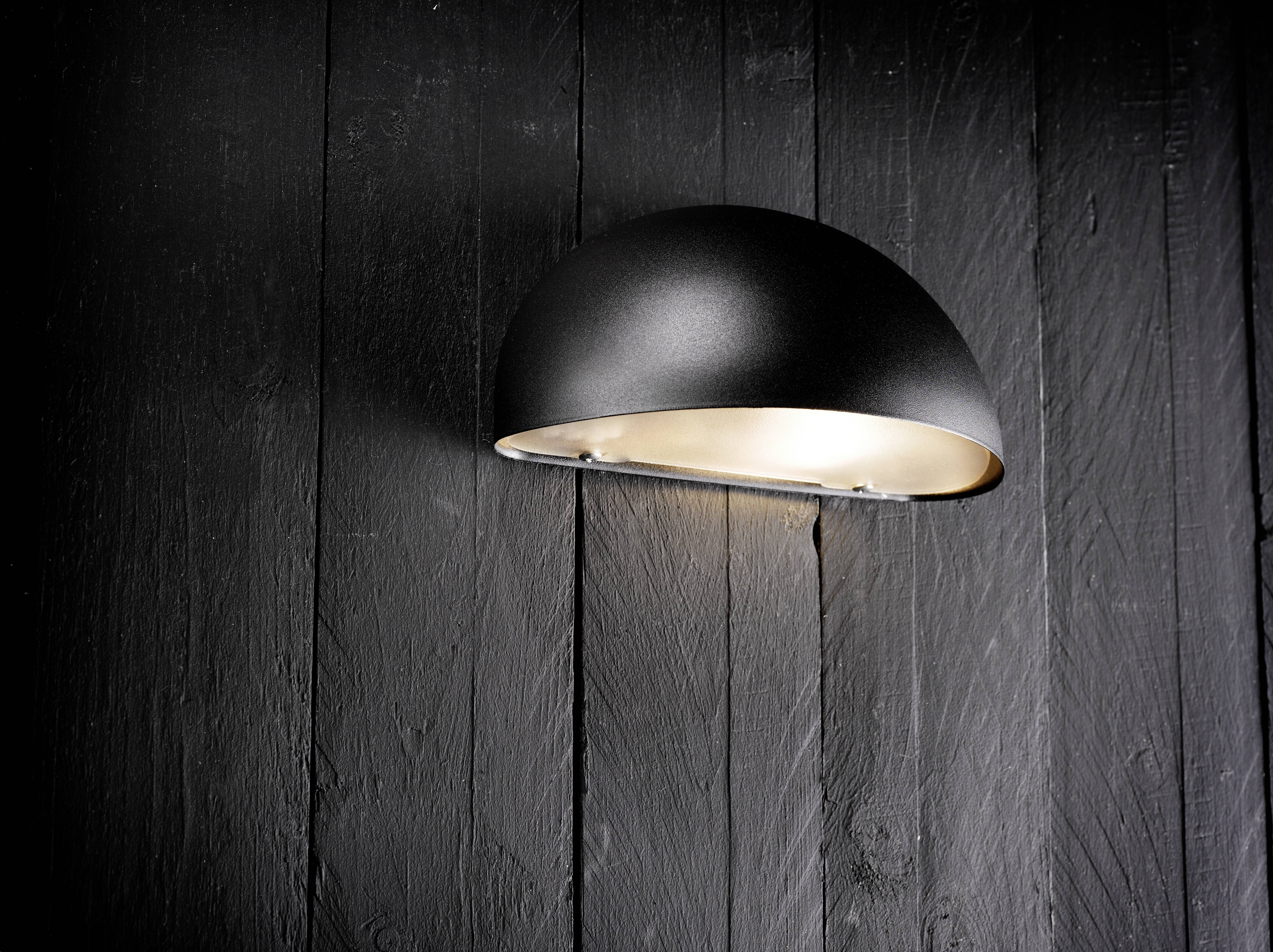 Ein schwarzes, kuppelförmiges Wandlicht auf einer dunklen Holzbretter-Wand montiert, das ein sanftes Licht nach unten wirft.