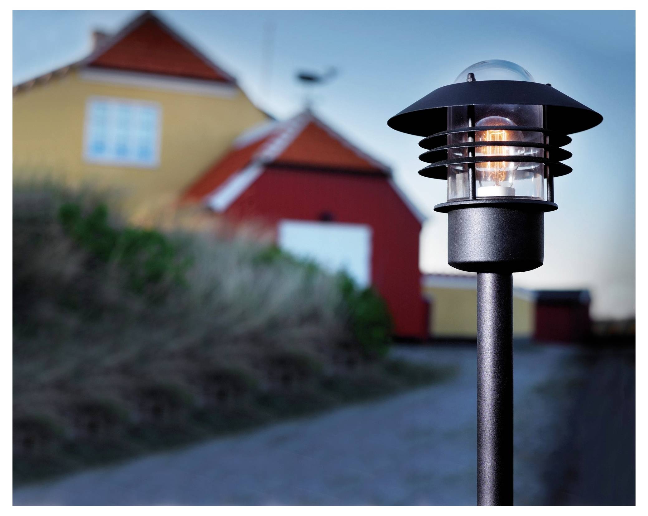 Ein beleuchteter Außenlampenpfahl steht im Vordergrund mit unscharfen gelben und roten Häusern im Hintergrund, wodurch eine gemütliche, dämmrige Atmosphäre entsteht.