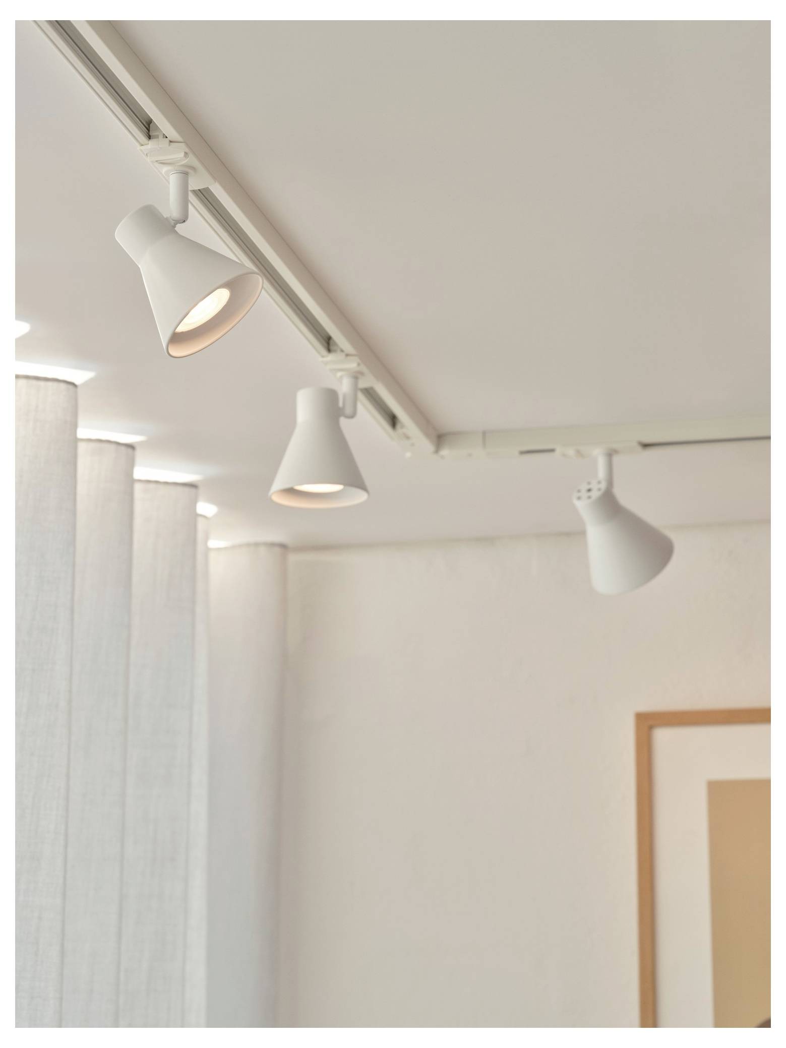 Deckenleuchtsystem mit drei weißen verstellbaren Strahlern, die einen modernen, minimalistischen Innenraum mit weißen Wänden und einem gerahmten Bild beleuchten.