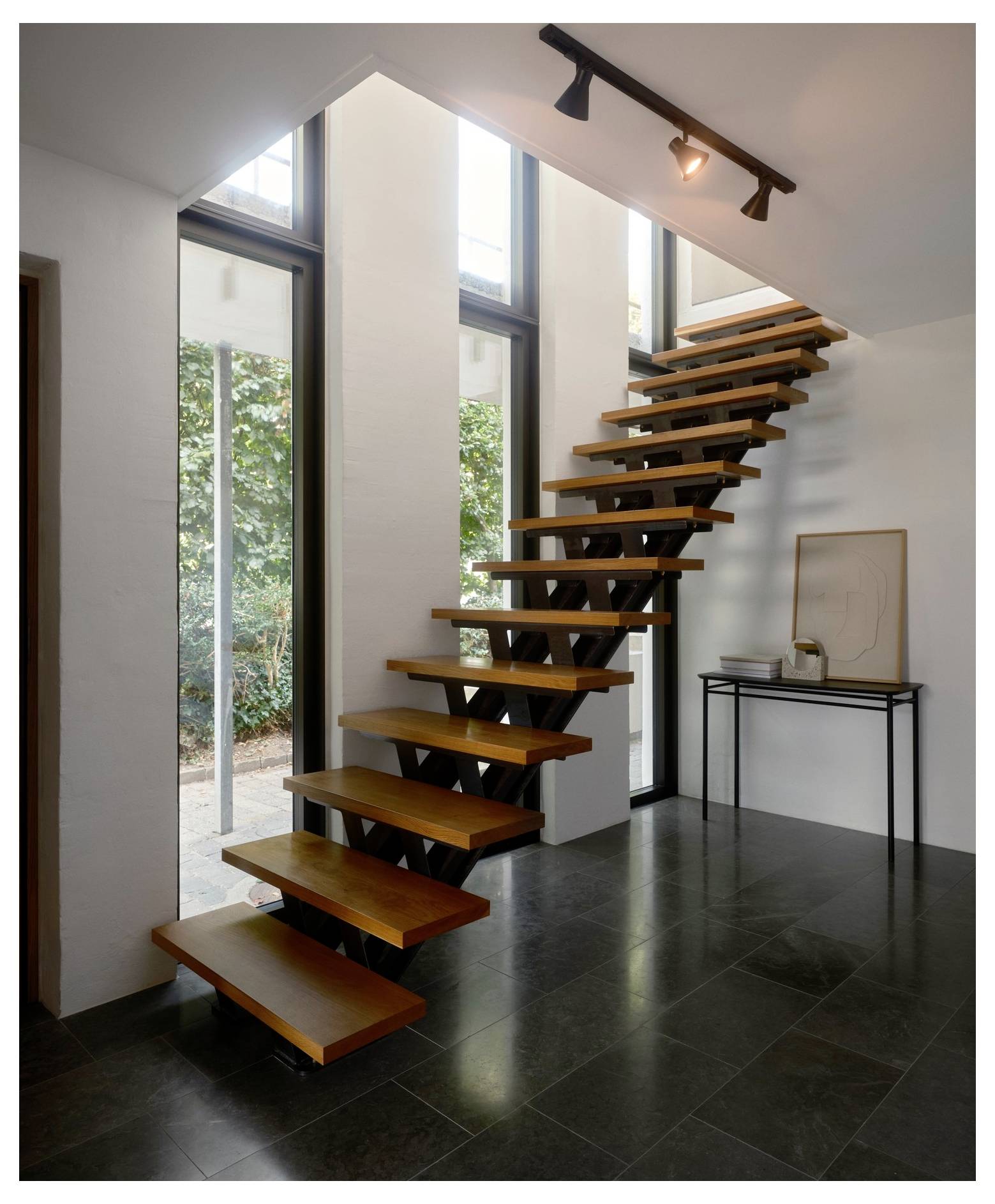 Eine moderne Holztreppe mit offenen Setzstufen führt in einem hellen Raum mit großen Fenstern nach oben. Ein kleiner Tisch darunter hält Kunstwerk und Dekoration.