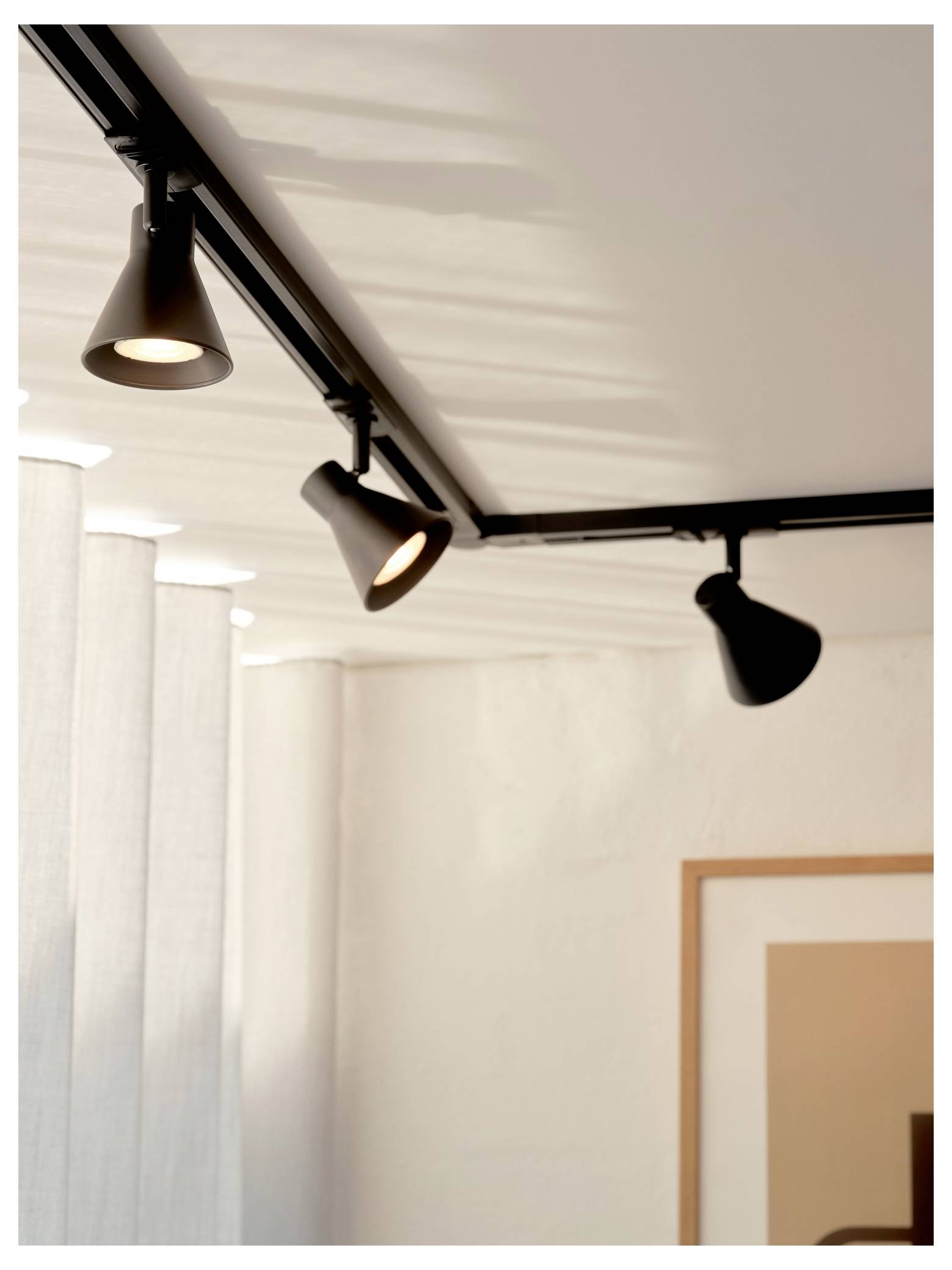 Schienensystem mit drei verstellbaren schwarzen Spotlights an einer weißen Decke, die einen minimalistischen Raum mit hellgrauen Vorhängen beleuchten.