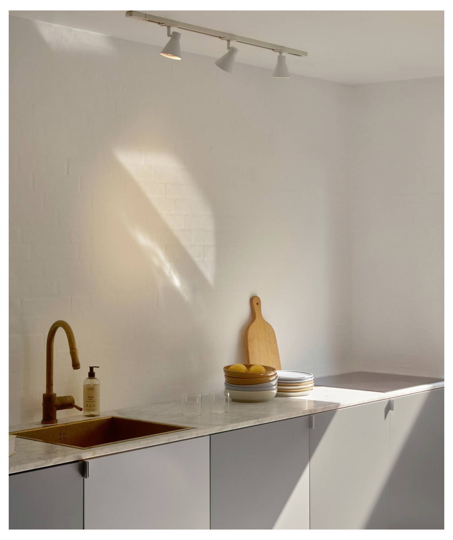 Eine minimalistische Küche mit grauen Schränken, einem Holzschneidebrett, einer Obstschale und einer Messingarmatur. Sonnenlicht wirft einen warmen Glanz.