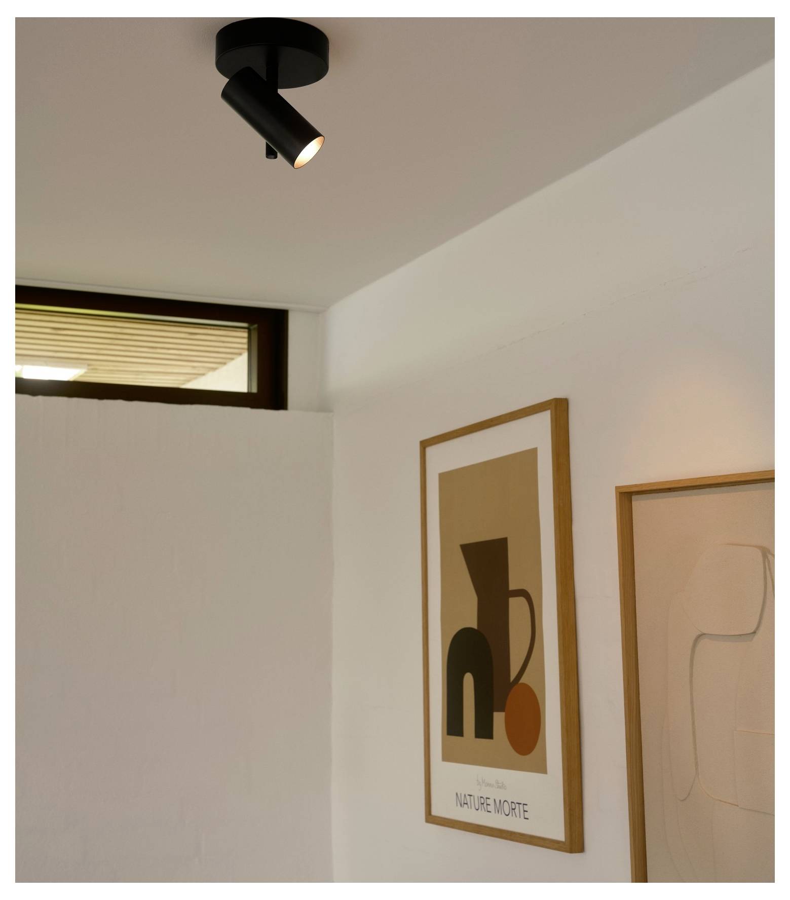 Ein moderner Deckenspot beleuchtet ein minimalistisches Zimmer mit abstrakter Wandkunst. Natürliches Licht fällt durch ein hohes Fenster auf der linken Seite ein.