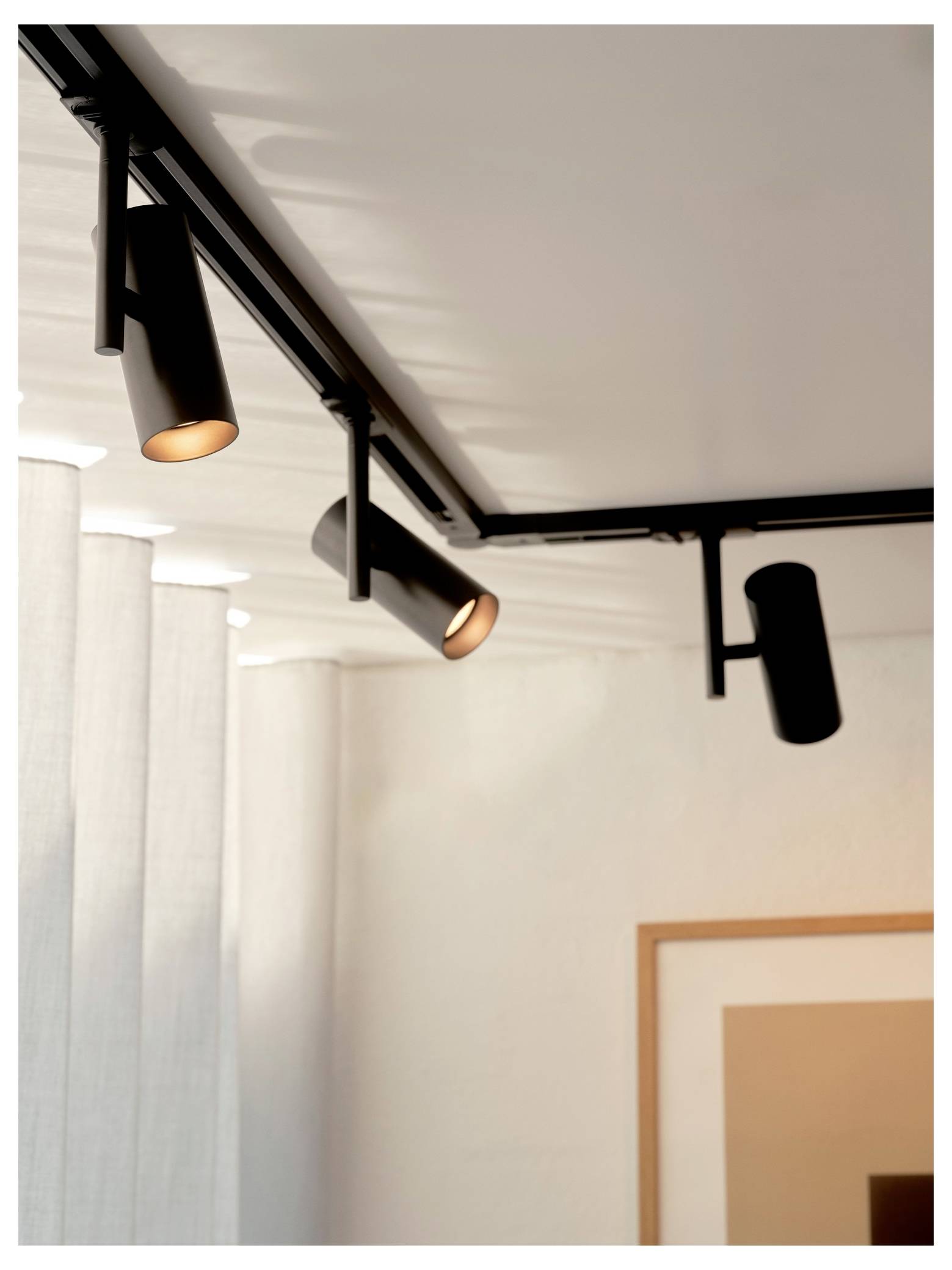 Drei schwarze Schienenstrahler sind an einer Decke montiert und beleuchten eine weiße Wand mit einem teilweise sichtbaren Rahmenbild im Hintergrund.