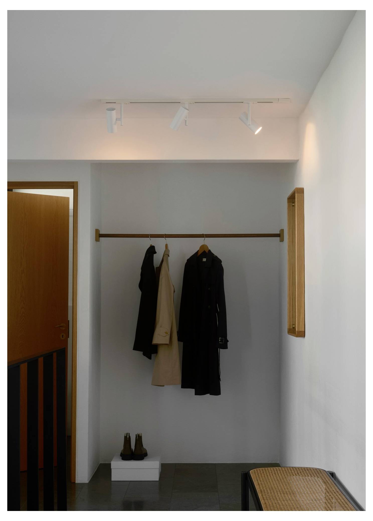 Ein minimalistischer Flur mit einer hölzernen Garderobe, die zwei Mäntel hält. Ein Paar Schuhe ist ordentlich auf einer kleinen weißen Plattform darunter platziert.