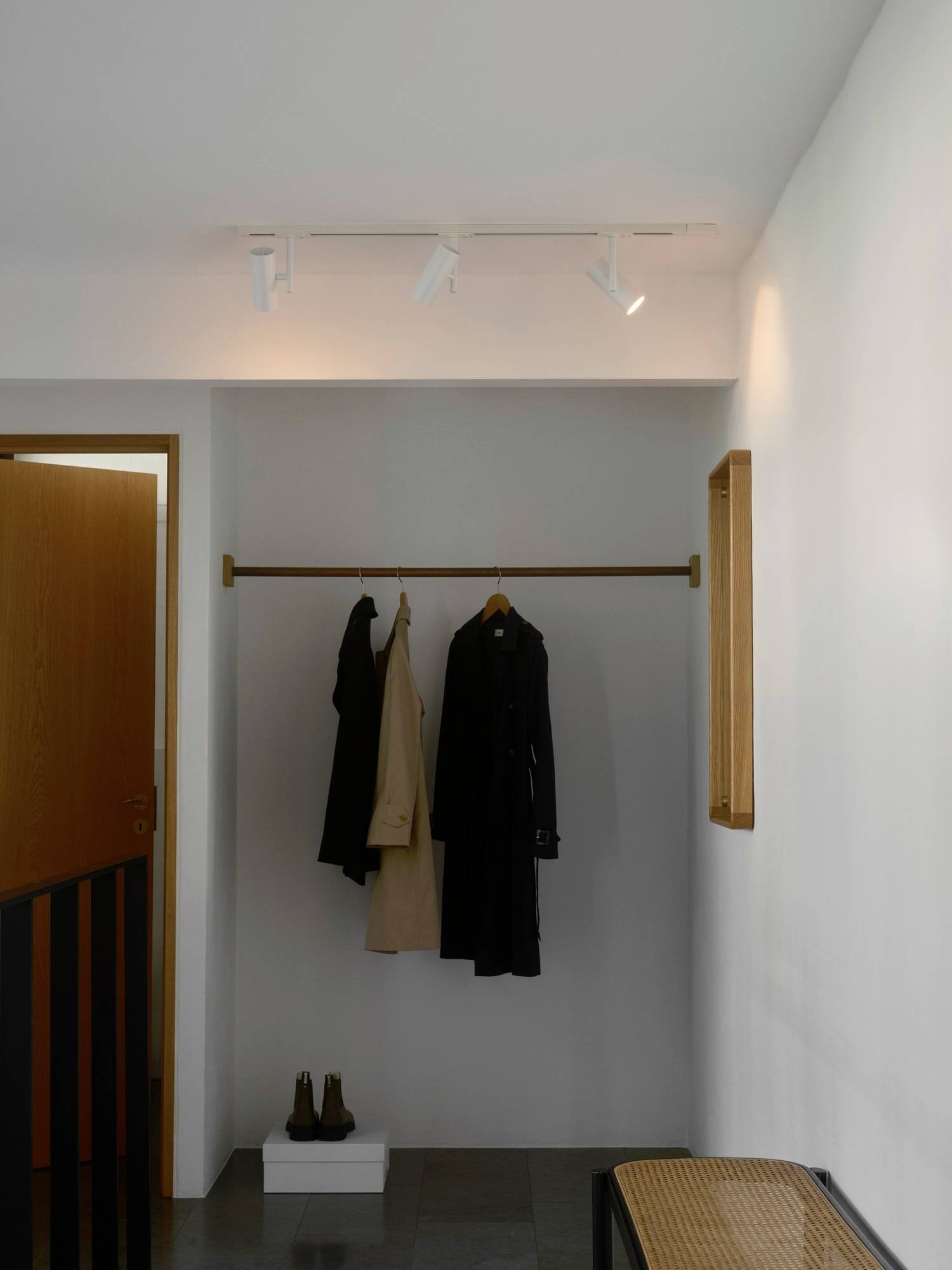 Ein minimalistischer Flur mit einer hölzernen Garderobe, auf der zwei Mäntel hängen. Darunter stehen ein Paar Stiefel auf dem Boden, mit sanfter Deckenbeleuchtung.