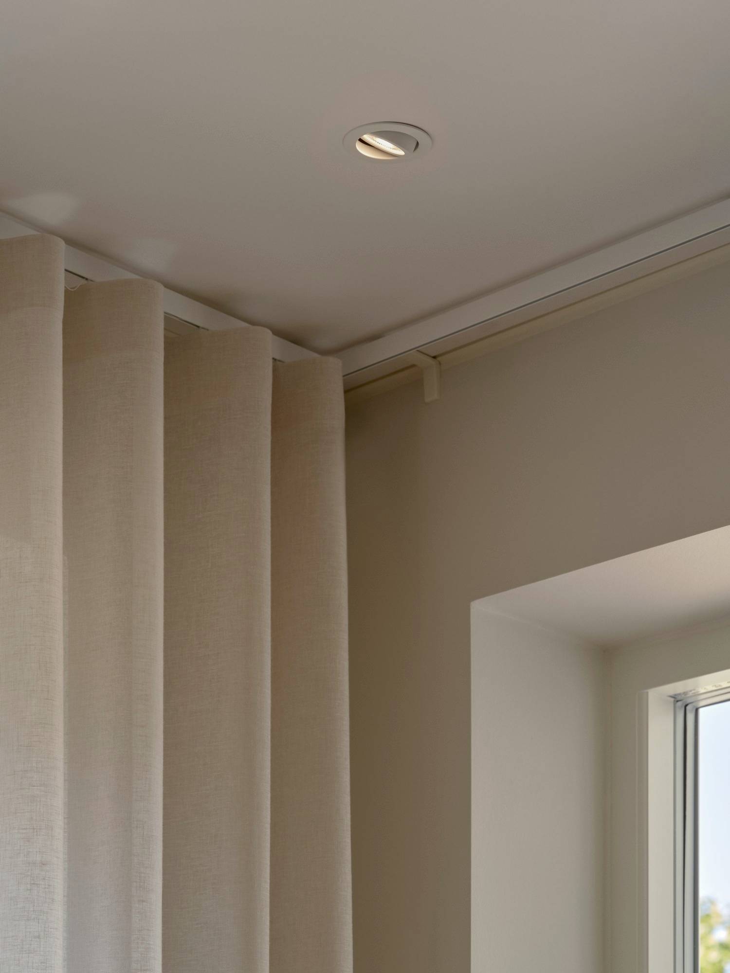 Decke mit eingelassener Leuchte über beigefarbenen Vorhängen in der Nähe eines teilweise sichtbaren Fensters, das eine weiche, stimmungsvolle Raumatmosphäre schafft.