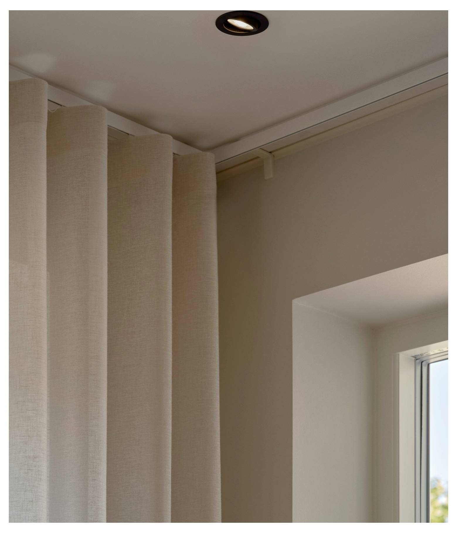 Ein an der Decke montierter Scheinwerfer beleuchtet beige Vorhänge neben einem Fenster und schafft eine warme, einladende Atmosphäre in einem modernen Innenraum.