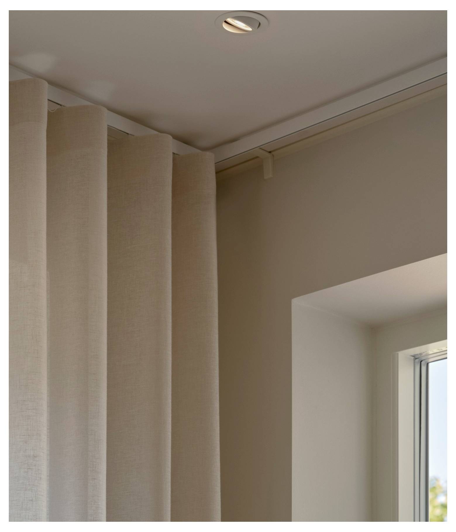 Eine Nahaufnahme von beigen Gardinen, die an einer an der Decke befestigten Schiene hängen, neben einem Fenster und schaffen eine weiche, elegante Innenraumatmosphäre.