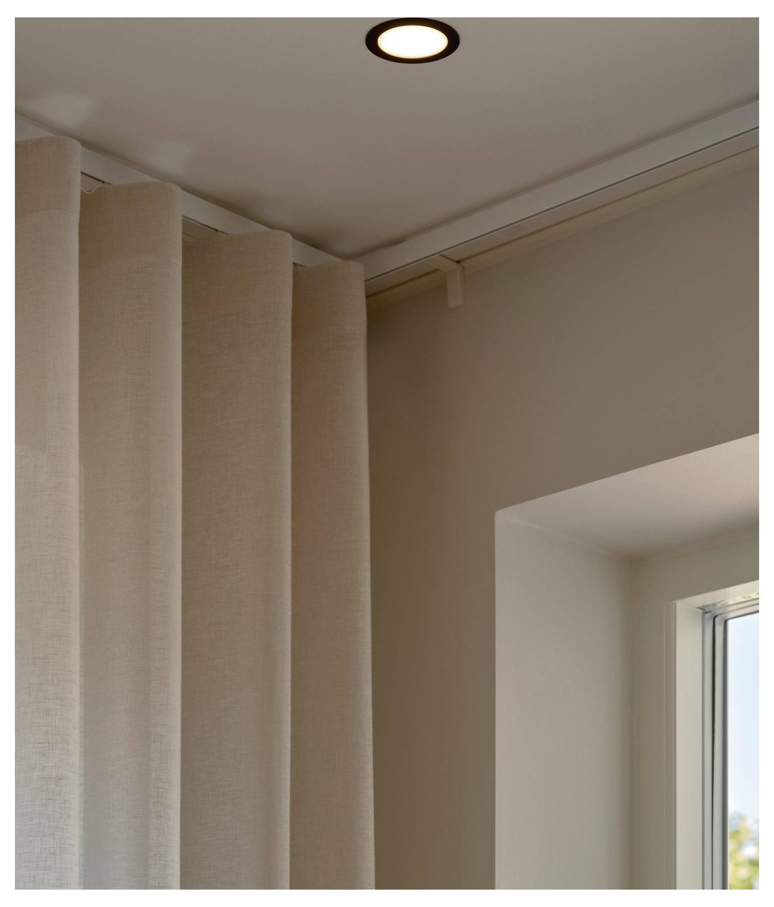 Eine Ecke eines Zimmers, die beige Vorhänge, eine Deckenleuchte und einen Teil eines Fensters mit Blick auf den Himmel zeigt.