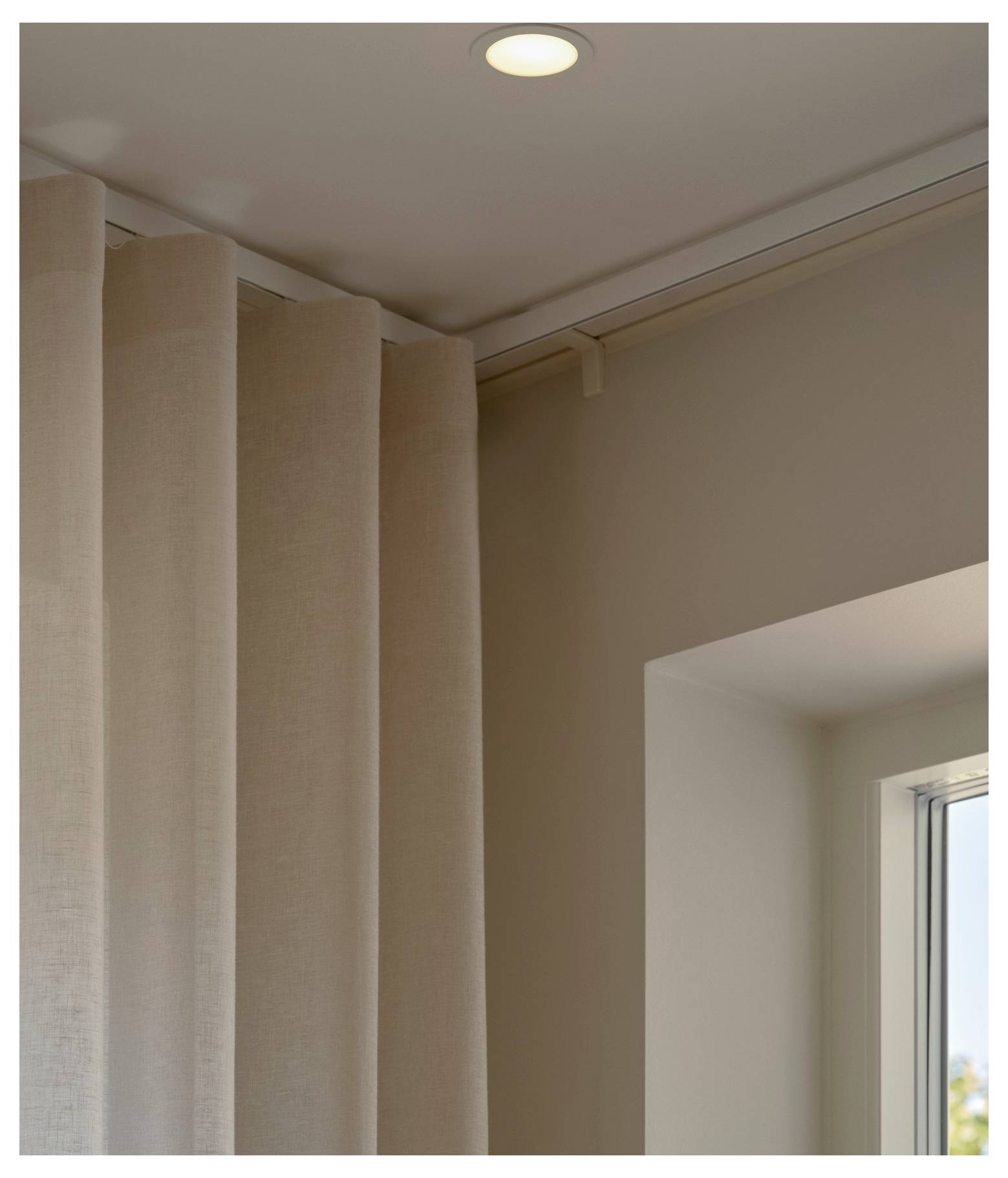 Ein beiger Vorhang hängt in der Ecke eines Zimmers neben einem Fenster, wobei eine Deckenleuchte eingeschaltet ist und eine gemütliche Atmosphäre schafft.