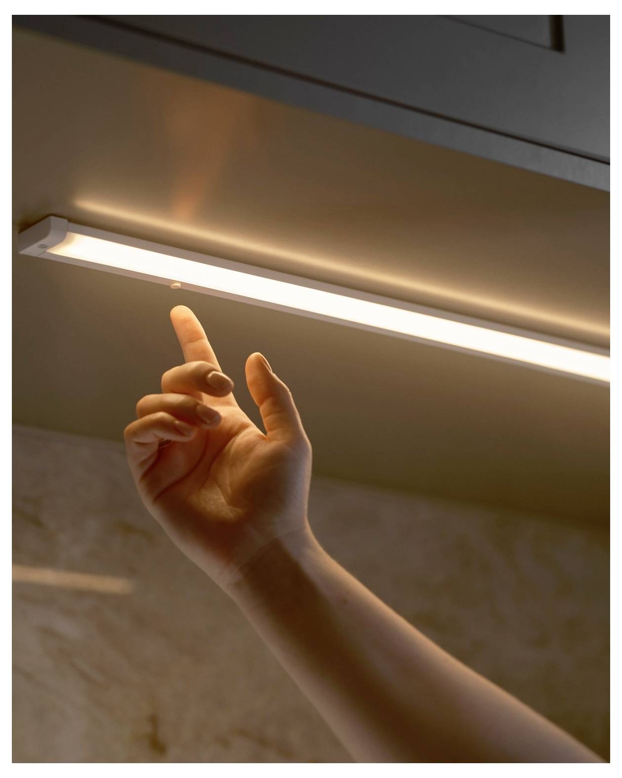 Eine Hand greift nach oben, um ein Lichtband unter einem Schrank einzuschalten und beleuchtet dabei eine Küchenarbeitsplatte.