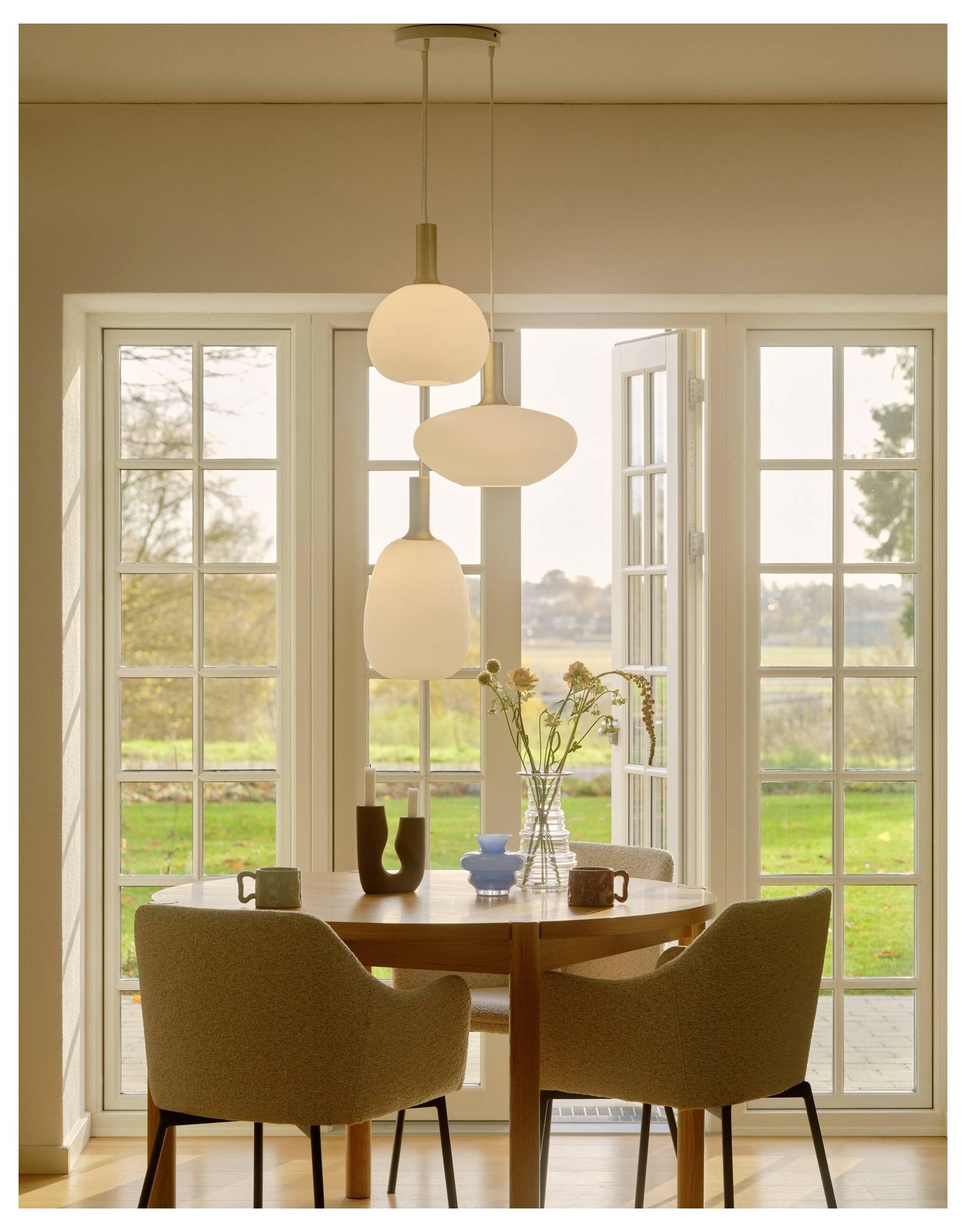 Ein gemütlicher Essbereich mit einem runden Tisch, zwei beigen Stühlen, modernen Pendelleuchten und einem Fensterblick auf einen grünen Garten draußen.