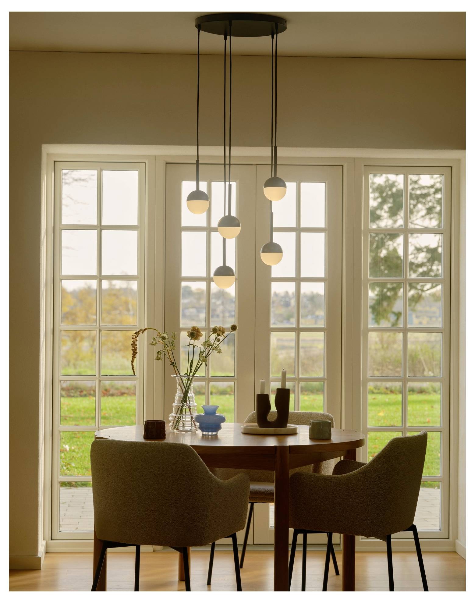 Ein gemütlicher Essbereich verfügt über einen runden Holztisch, zwei beige Stühle, Pendelleuchten und eine Vase mit Blumen, die sich vor großen Glastüren mit Blick auf einen Garten befindet.