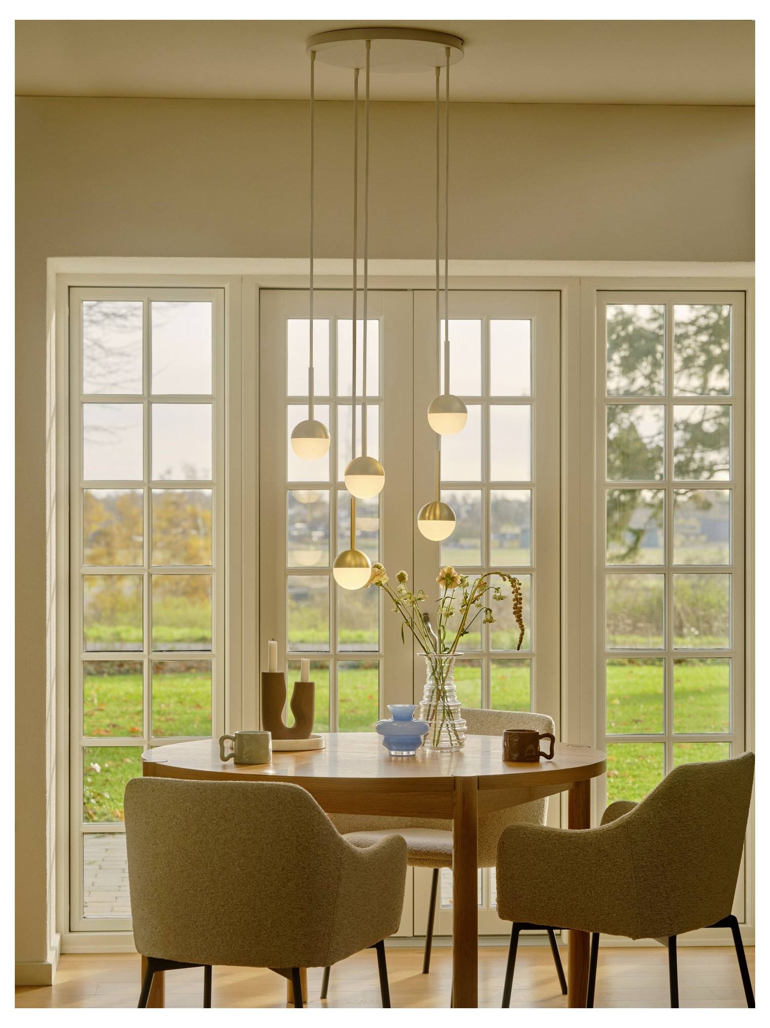 Ein runder Holztisch mit zwei gepolsterten Stühlen vor großen Fenstern mit Gartenblick. Pendelleuchten hängen darüber, und eine Vase mit Blumen steht auf dem Tisch.