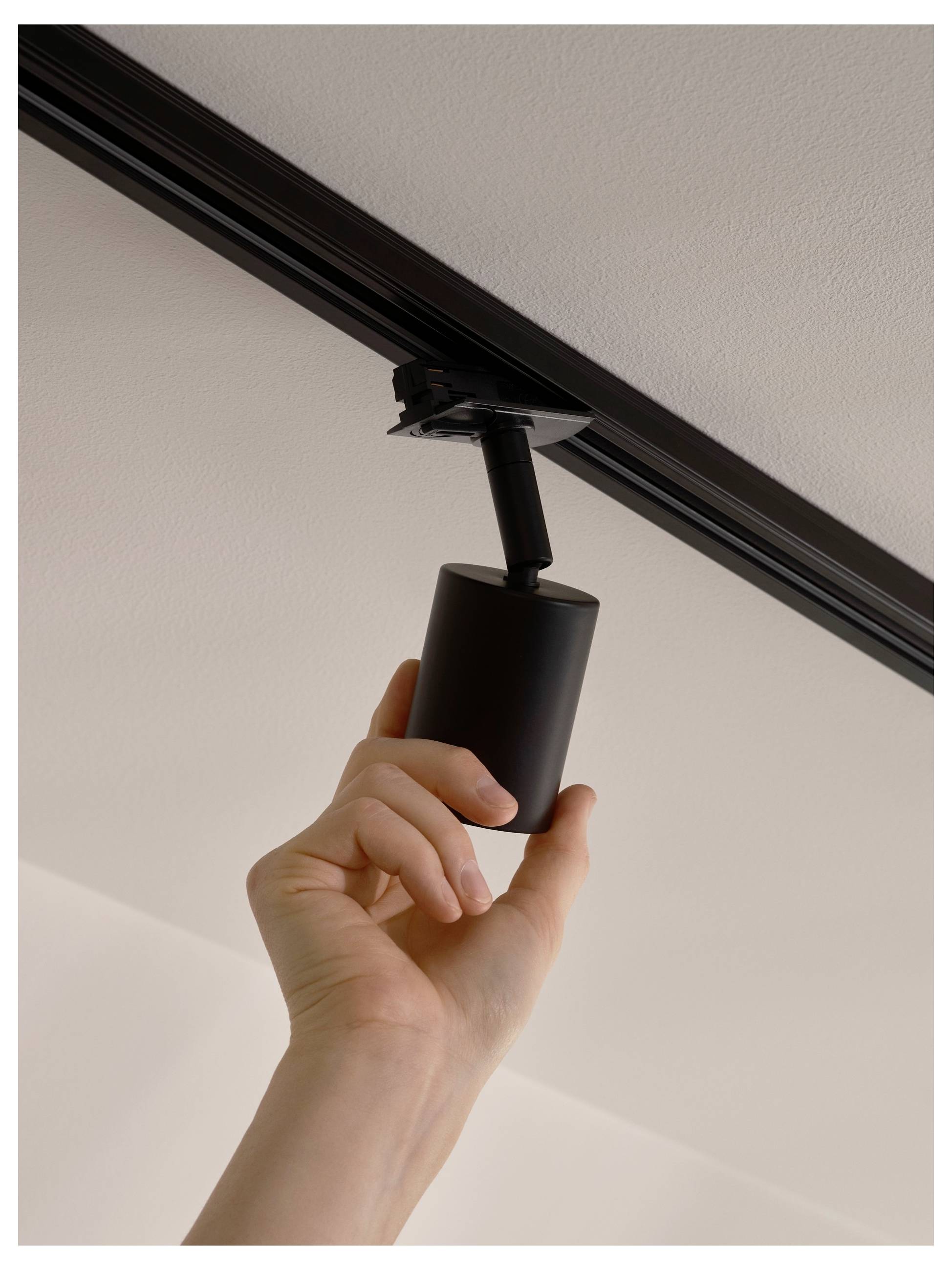 Eine Hand installiert oder passt einen schwarzen, an der Decke montierten Strahler an, wobei der Fokus auf der Beleuchtungseinrichtung im Haus oder Büro liegt.
