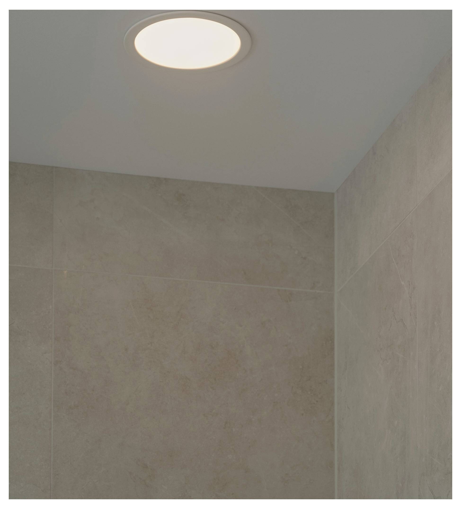 Eine eingelassene Deckenleuchte beleuchtet eine Ecke eines Raums mit beigefarbenen gekachelten Wänden und hebt dabei die glatte Textur und neutralen Töne hervor.