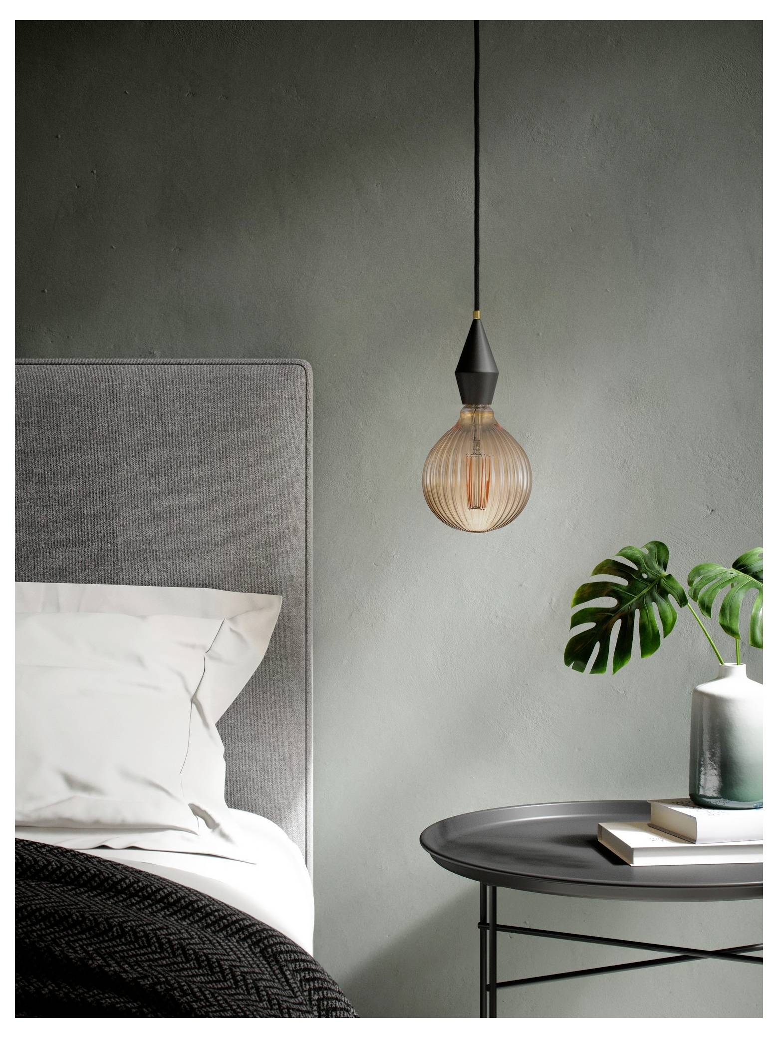 Eine moderne Schlafzimmerszene mit einer hängenden Pendelleuchte, einem grau gepolsterten Bett mit weißer Bettwäsche und einem kleinen Tisch mit einer Pflanze und Büchern.