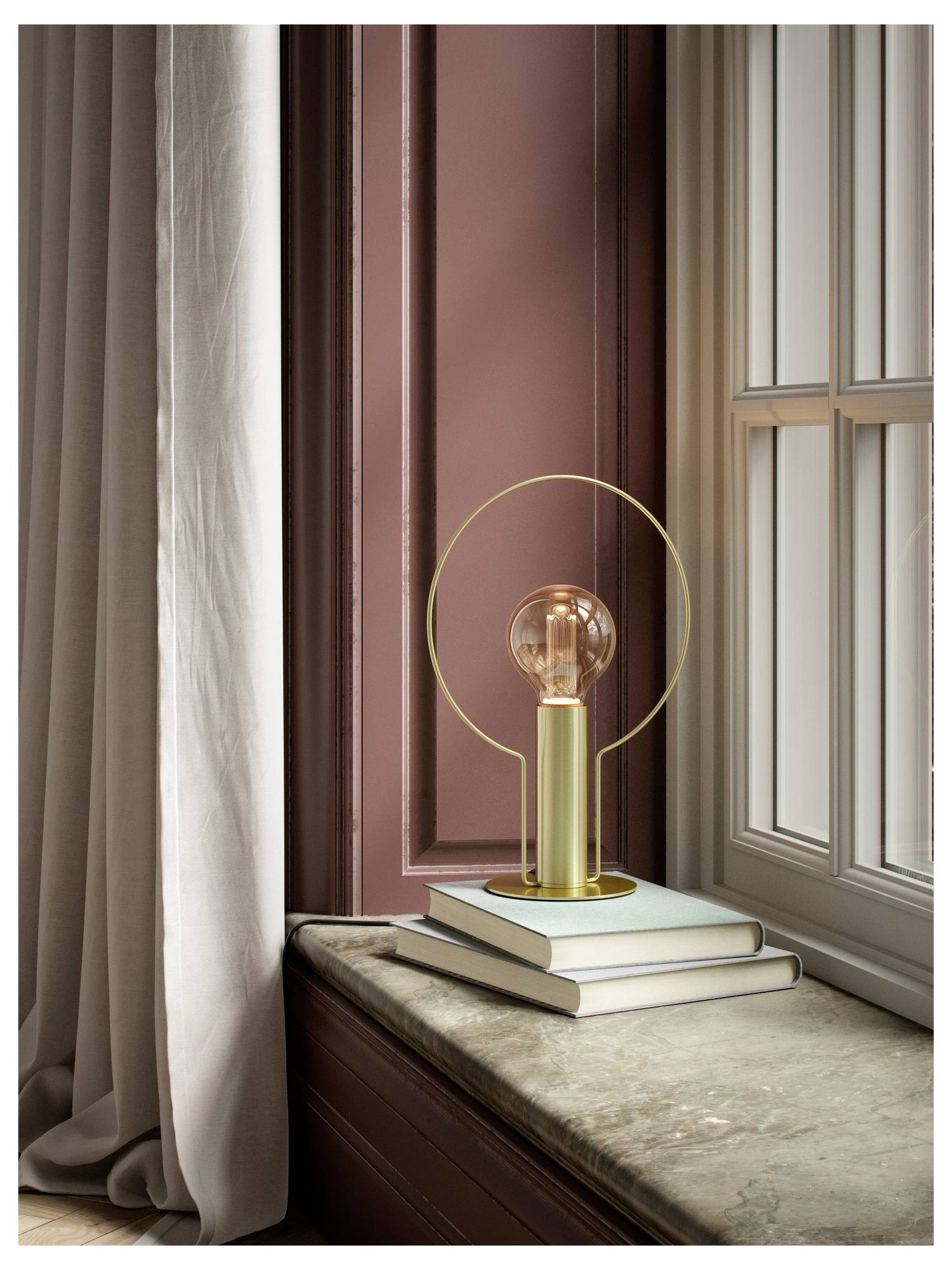 Eine moderne Lampe mit einem kreisförmigen Metallrahmen und einer freiliegenden Glühbirne sitzt auf Büchern neben einem Fenster mit rosa Paneelen und bodenlangetvorhängen.