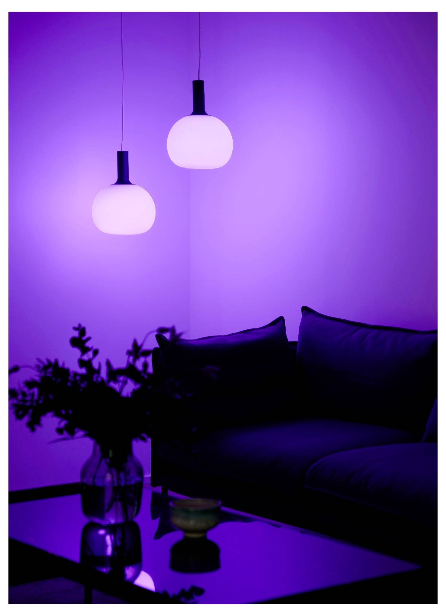 Ein schwach beleuchteter Raum mit zwei leuchtenden Pendelleuchten und einem Sofa. Ein Glastisch mit einer Blumenvase und einer kleinen Schale trägt zu der gemütlichen Atmosphäre bei.