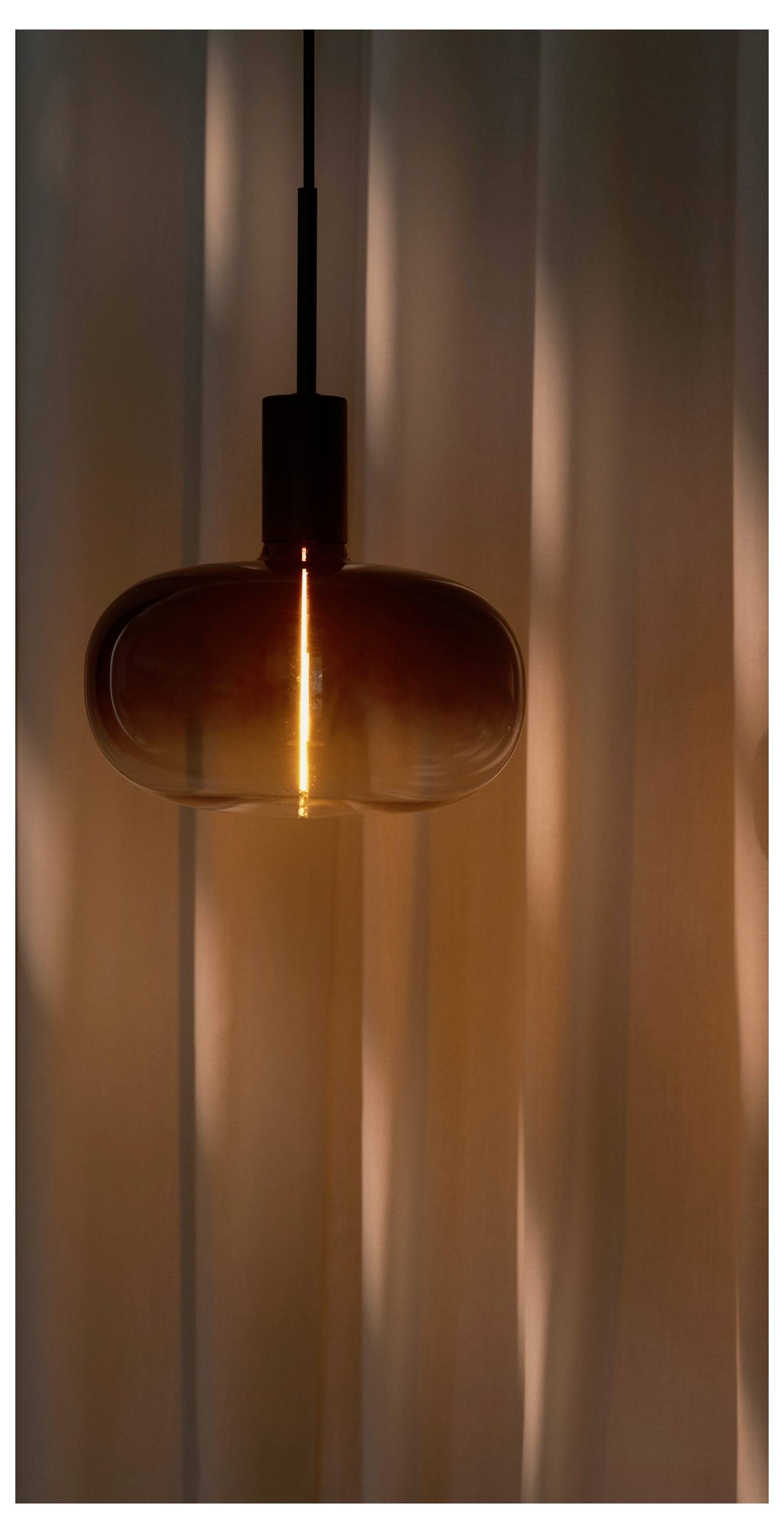 Eine warme, leuchtende Pendelleuchte mit rundem Glassschirm hängt vor sanft beleuchteten Vorhängen und schafft eine gemütliche und einladende Atmosphäre.