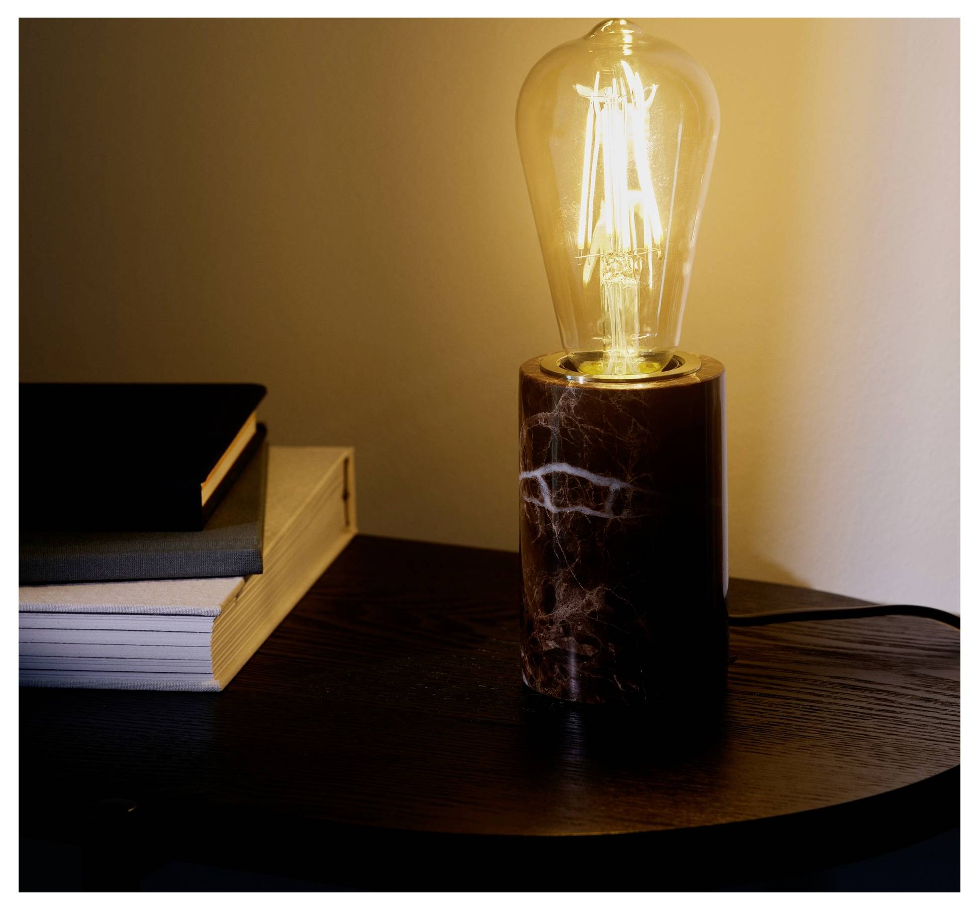 Eine beleuchtete Glühbirne im Vintage-Stil auf einem Marmorsockel sitzt auf einem dunklen Holztisch neben gestapelten Büchern und wirft ein warmes Licht.