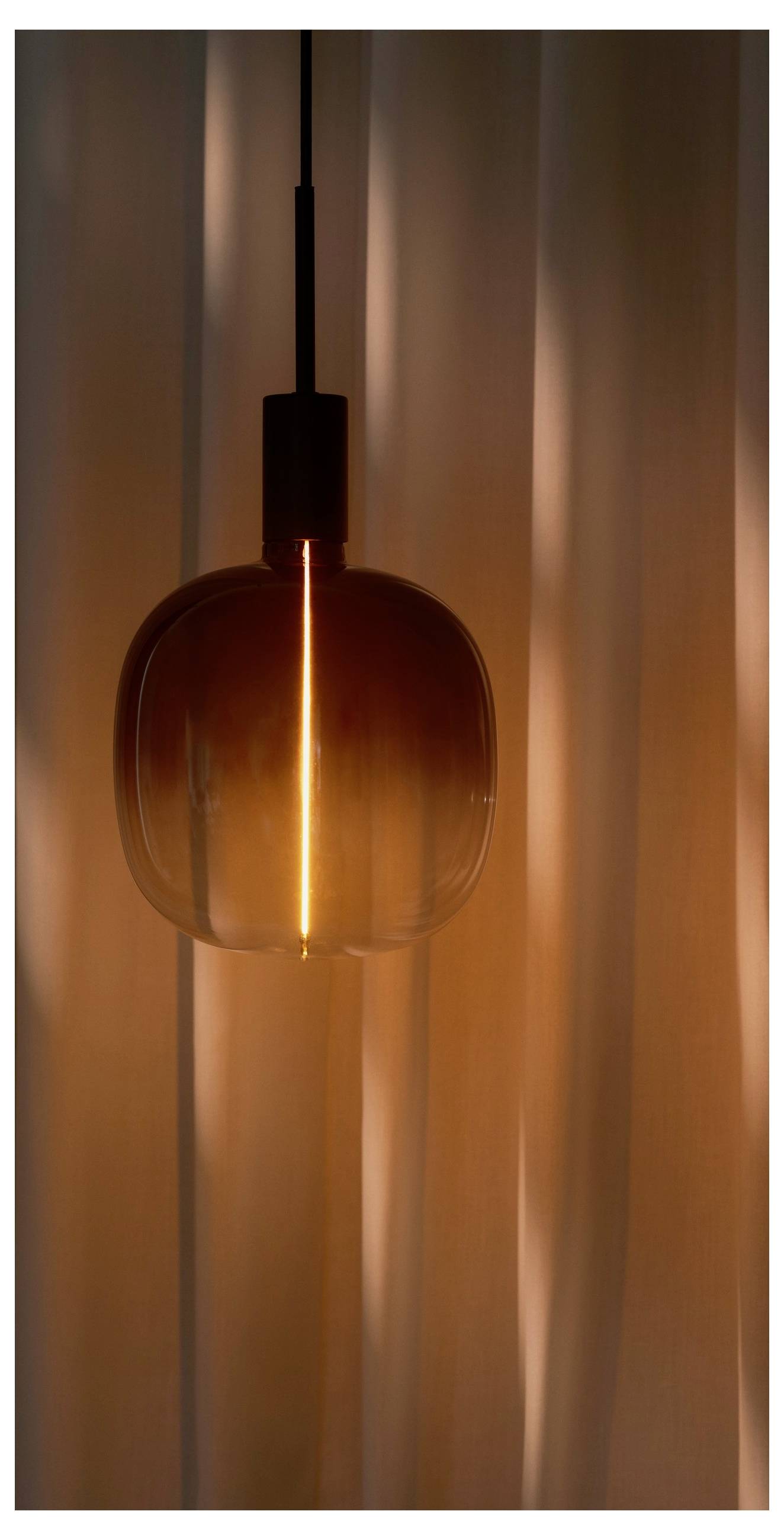 Eine warmtonige Pendelleuchte leuchtet vor einem sanft unscharfen Vorhanghintergrund und schafft eine gemütliche, stimmungsvolle Atmosphäre.