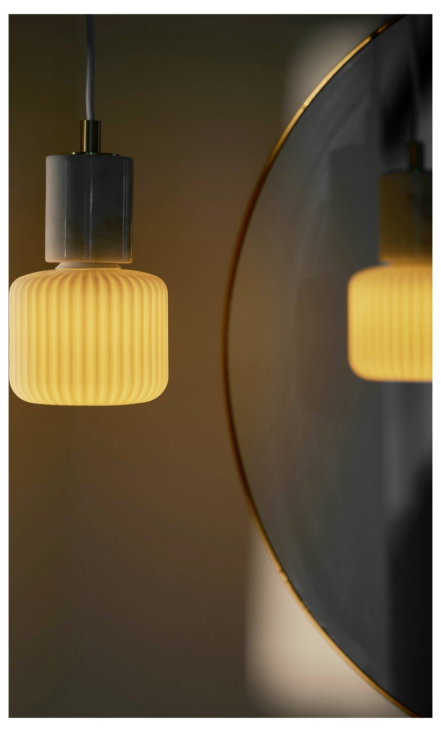 Eine einzelne gerippte Glaspendeleuchte mit warmem Glühen wird in einem nahegelegenen Spiegel reflektiert und erzeugt einen symmetrischen visuellen Effekt.