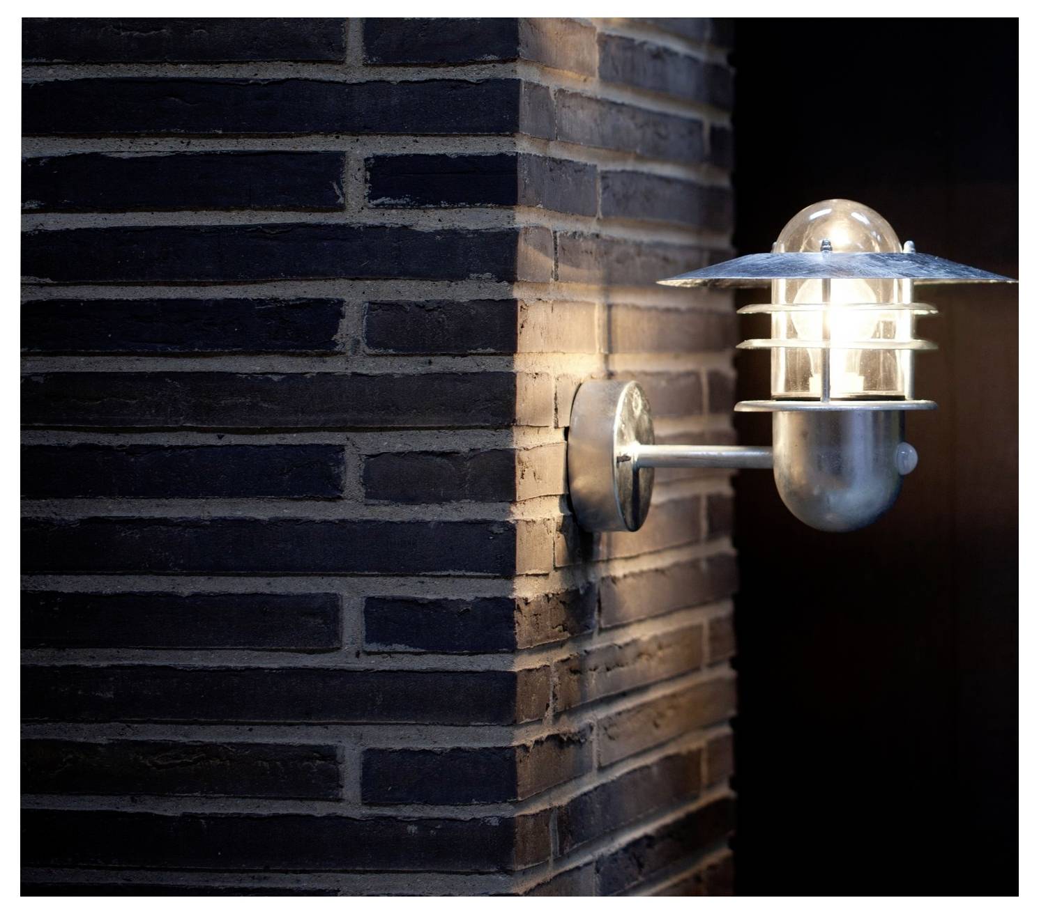 Eine wandmontierte Außenleuchte mit metallischem Rahmen beleuchtet eine Ecke mit dunklem Mauerwerk und einer glatten Oberfläche und wirft ein weiches Glühen.