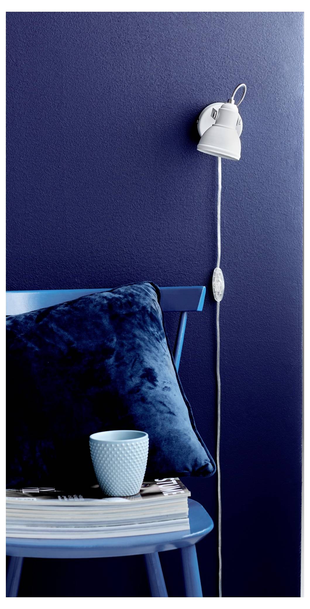 Ein blauer Stuhl mit einem dunkelblauem Kissen steht gegen eine marineblaue Wand. Ein Stapel Zeitschriften mit einer hellblauen Tasse ruht auf dem Stuhl. Eine Wandleuchte befindet sich darüber.