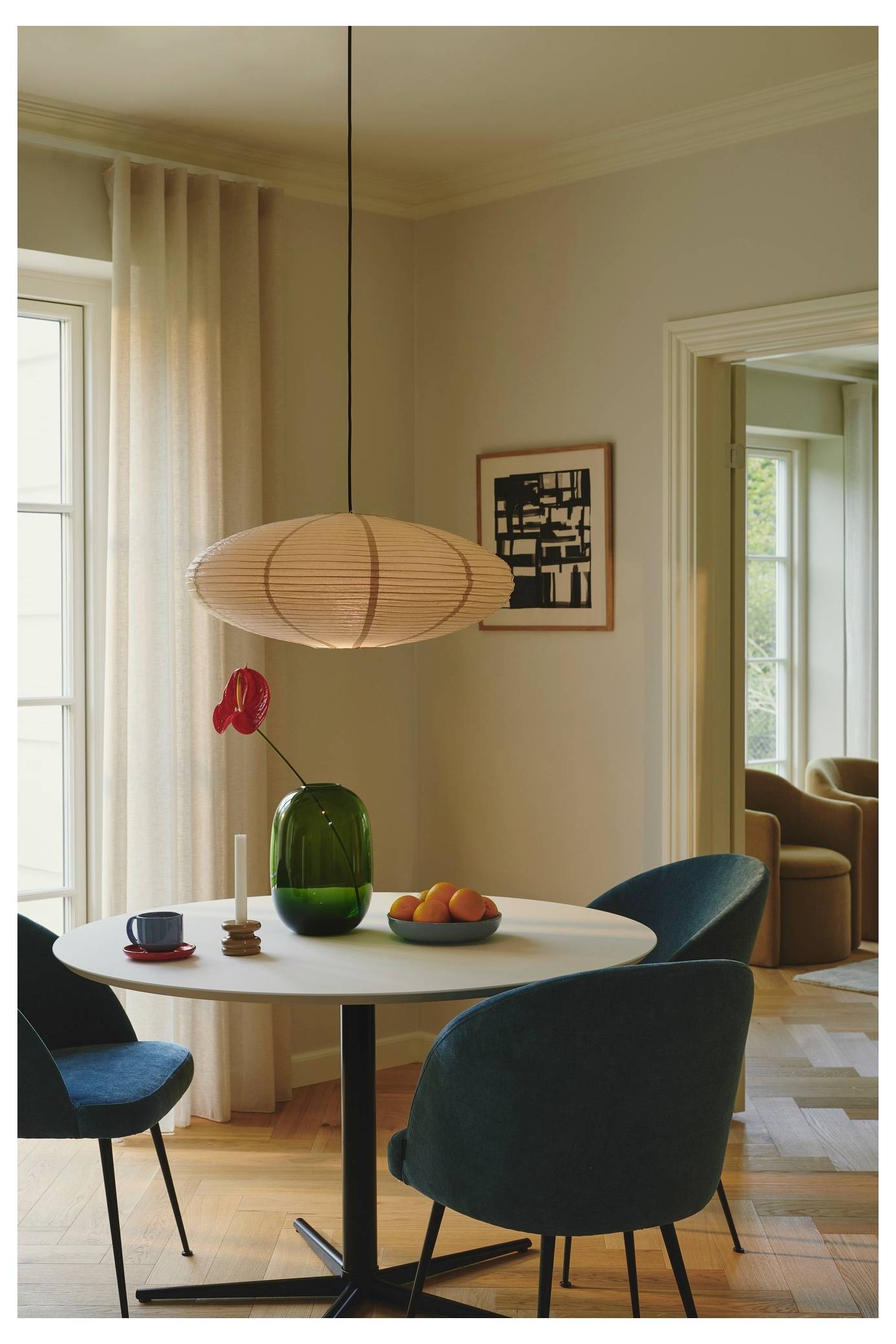 Ein moderner Essbereich mit einem runden weißen Tisch, türkisfarbenen Stühlen, einer grünen Vase mit roter Blume, einer Obstschale und einer Pendelleuchte; Sonnenlicht filtert durch halbdurchsichtige Vorhänge herein.