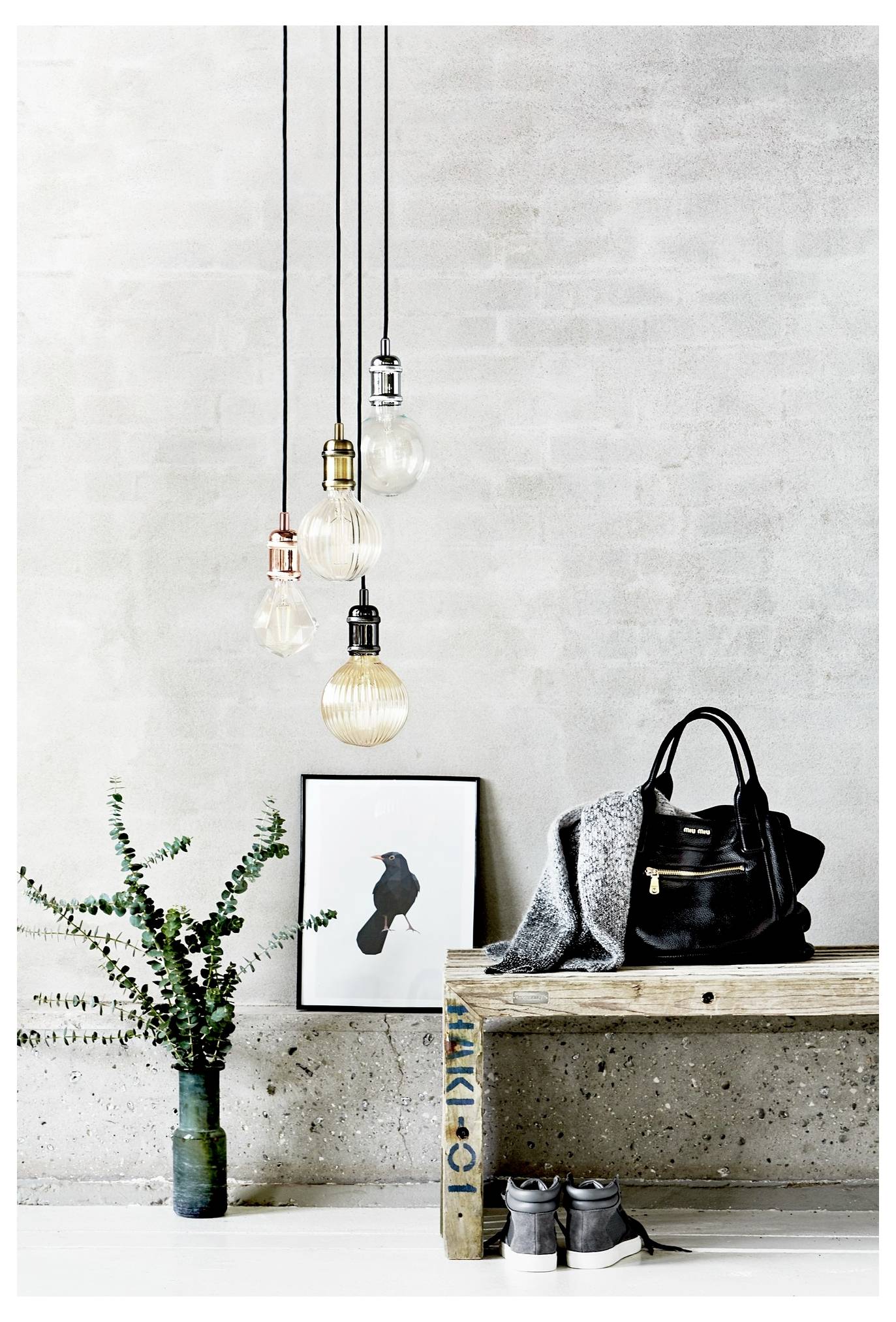 Ein minimalistisches Zimmer mit hängenden Vintage-Glühbirnen, einer Bank mit einer schwarzen Tasche und einem grauen Pullover, Sneakern, einem gerahmten Vogelkunstwerk und einer Vase mit Blattranken.