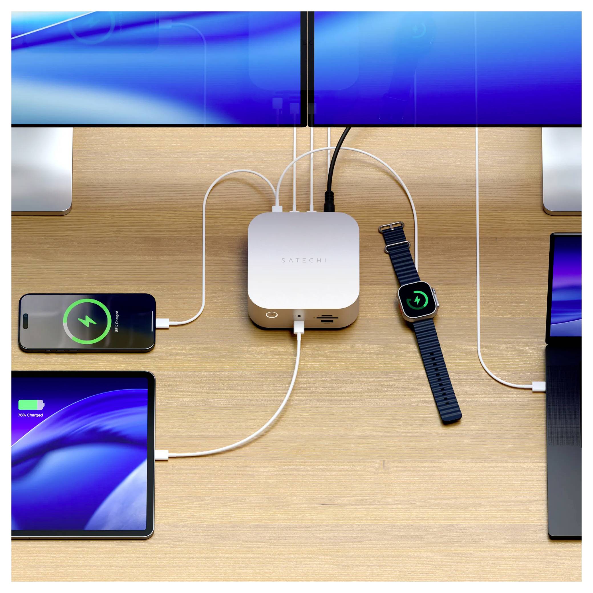 Draufsicht auf einen Schreibtisch mit einem Belkin-Ladegerät, das mit mehreren Geräten verbunden ist: zwei Telefone, ein Tablet, eine Smartwatch und Monitore, die alle über Kabel aufgeladen werden.