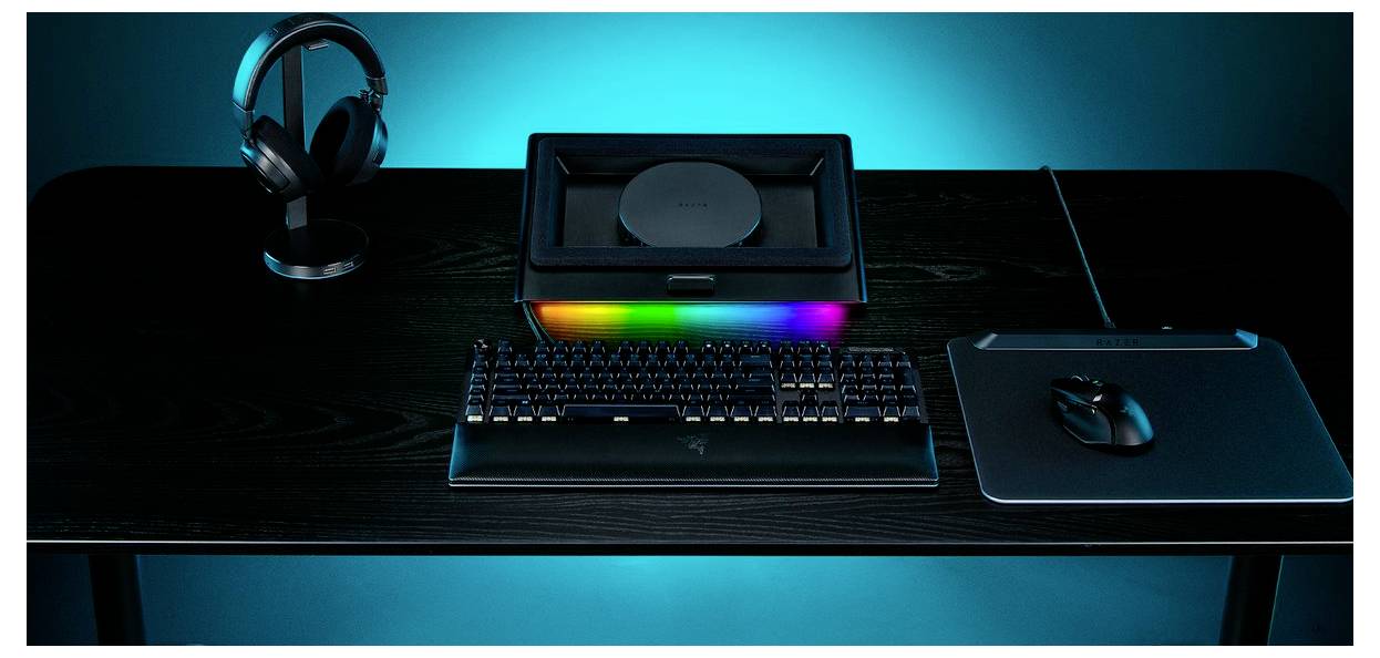 Ein eleganter schwarzer Schreibtisch zeigt eine Tastatur mit farbiger Hintergrundbeleuchtung, einen geschlossenen Laptop, Kopfhörer auf einem Ständer und eine Maus auf einem Mousepad.