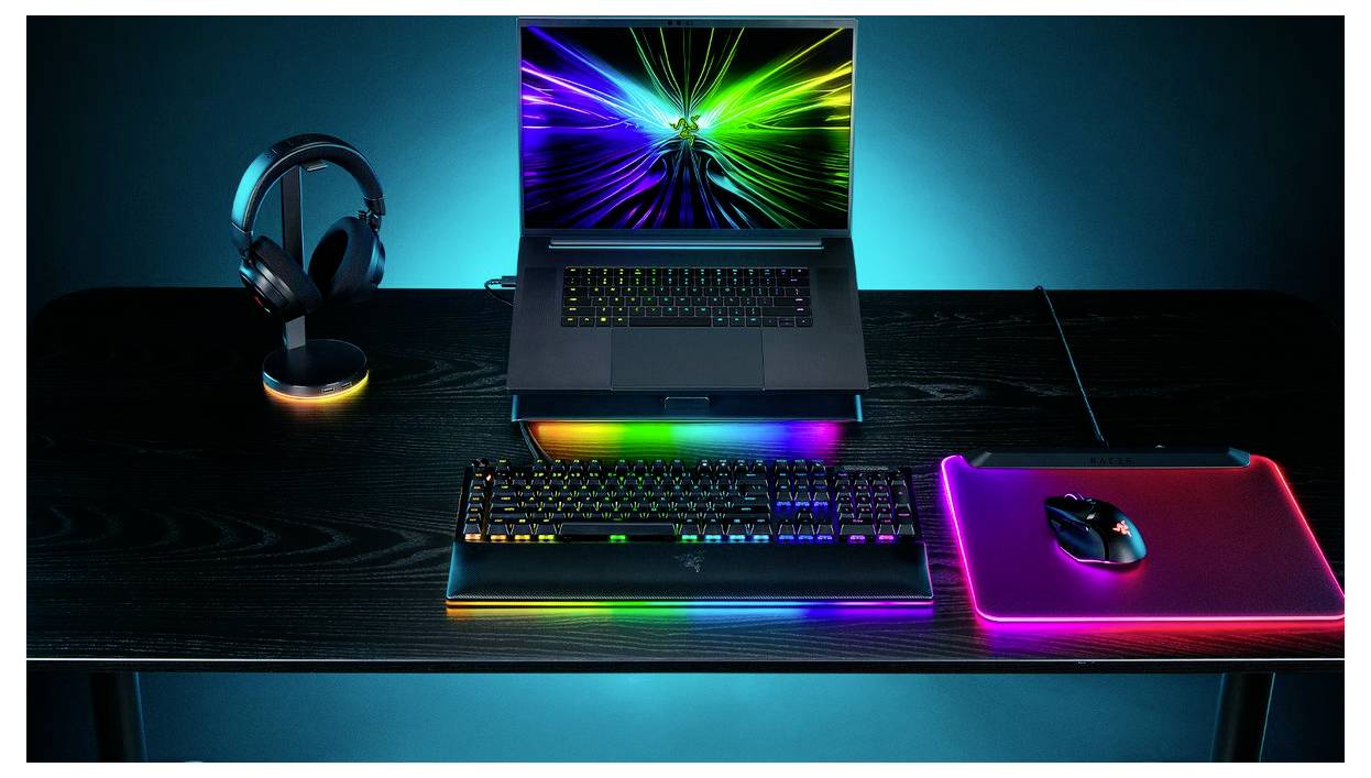 Ein farbenfroher Gaming-Setup mit einem Laptop, der ein Regenbogenmuster auf dem Bildschirm anzeigt, einer RGB-beleuchteten Tastatur, einer Maus auf einem beleuchteten Mousepad und Kopfhörern.