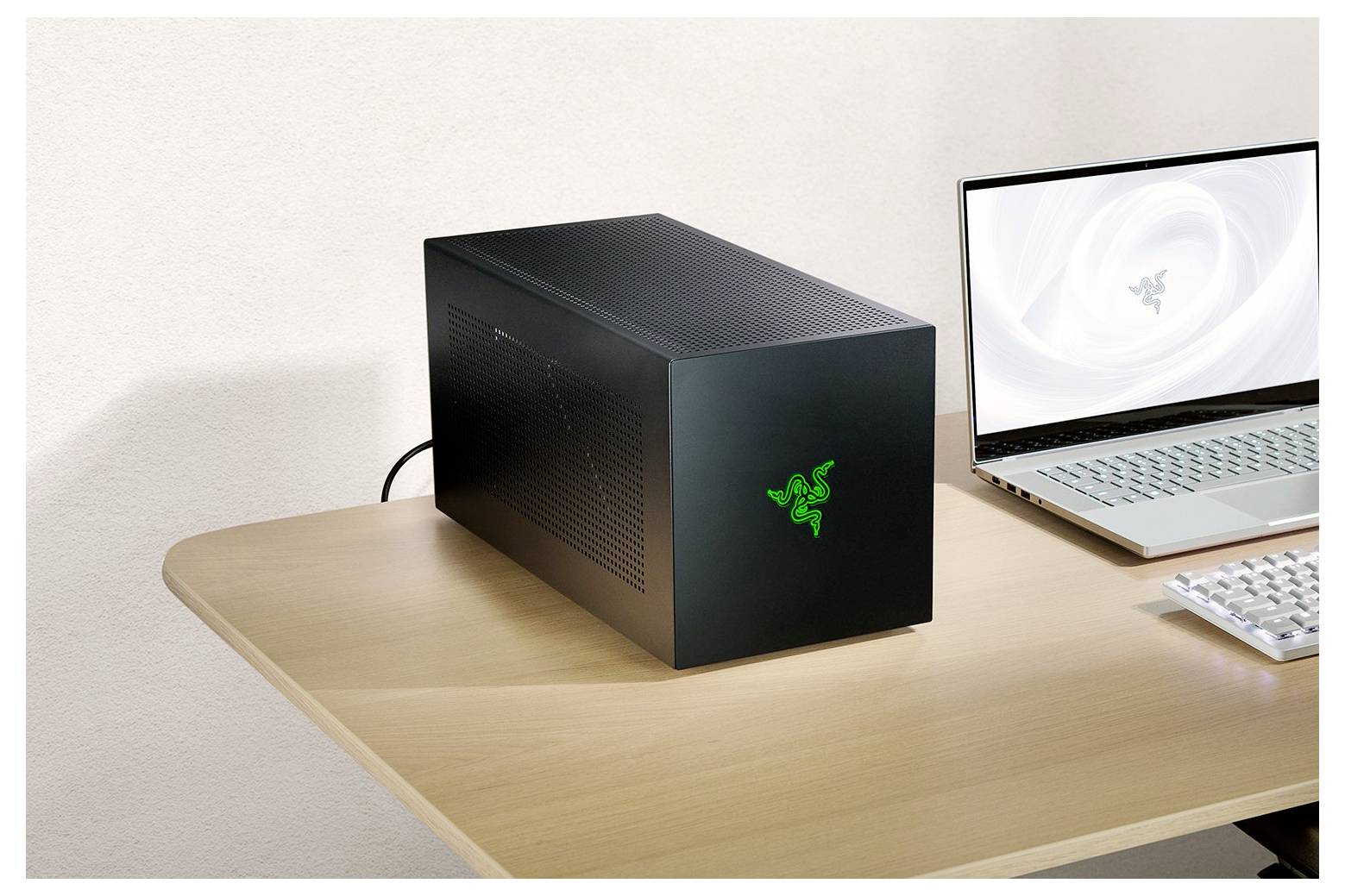 Ein schwarzes externes GPU-Gehäuse mit grünem Logo steht auf einem Holzschreibtisch neben einem aufgeklappten silbernen Laptop, der ein ähnliches Logo anzeigt.