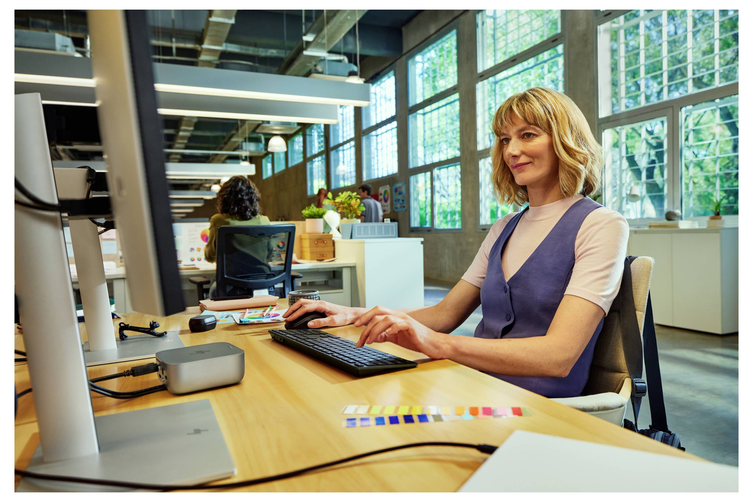 Eine Frau, die an einem Computer in einem modernen, hell beleuchteten Büro mit großen Fenstern arbeitet. Sie tippt auf einer Tastatur und wirkt konzentriert.