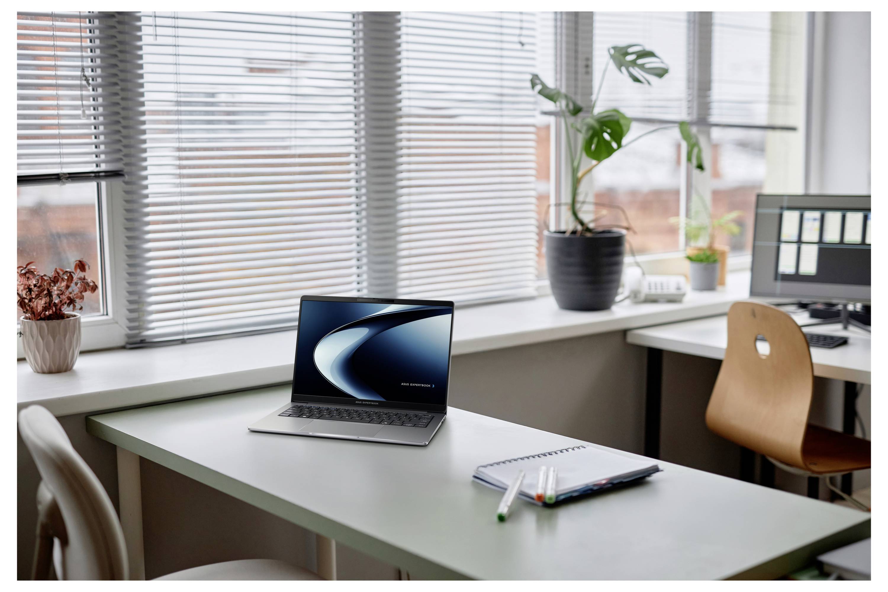 Ein moderner leerer Schreibtisch mit einem Laptop, einem Notizbuch und Stiften neben einem Fenster mit Jalousien, Pflanzen und einem Computermonitor.
