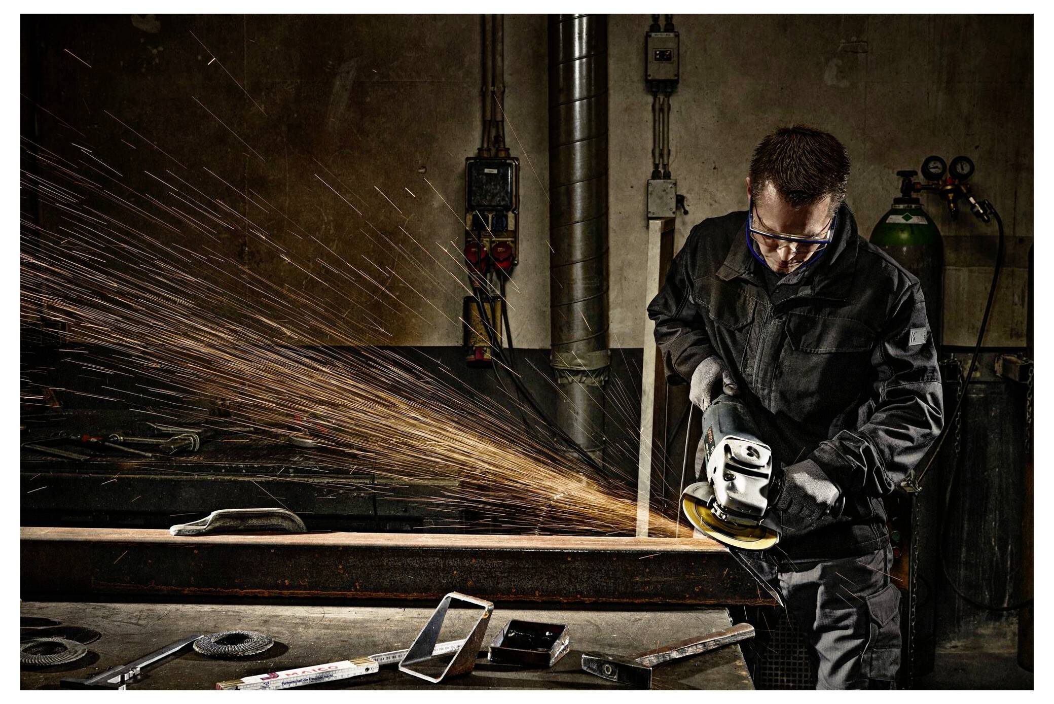 Ein Mann in einer Werkstatt benutzt einen Winkelschleiffer auf Metall und erzeugt dabei Funken. Er trägt eine Schutzbrille und Handschuhe und konzentriert sich auf die Aufgabe.