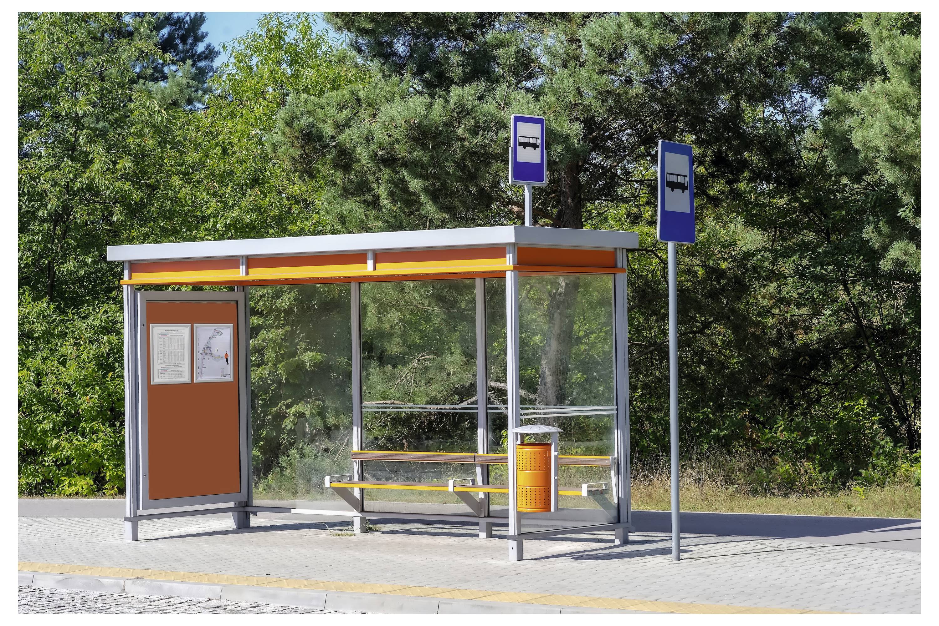 Ein kleines Bushaltestellen-Unterstand mit einer Bank, einer Karte und Schildern, das sich auf einer gepflasterten Fläche befindet und von Bäumen im Hintergrund umgeben ist.