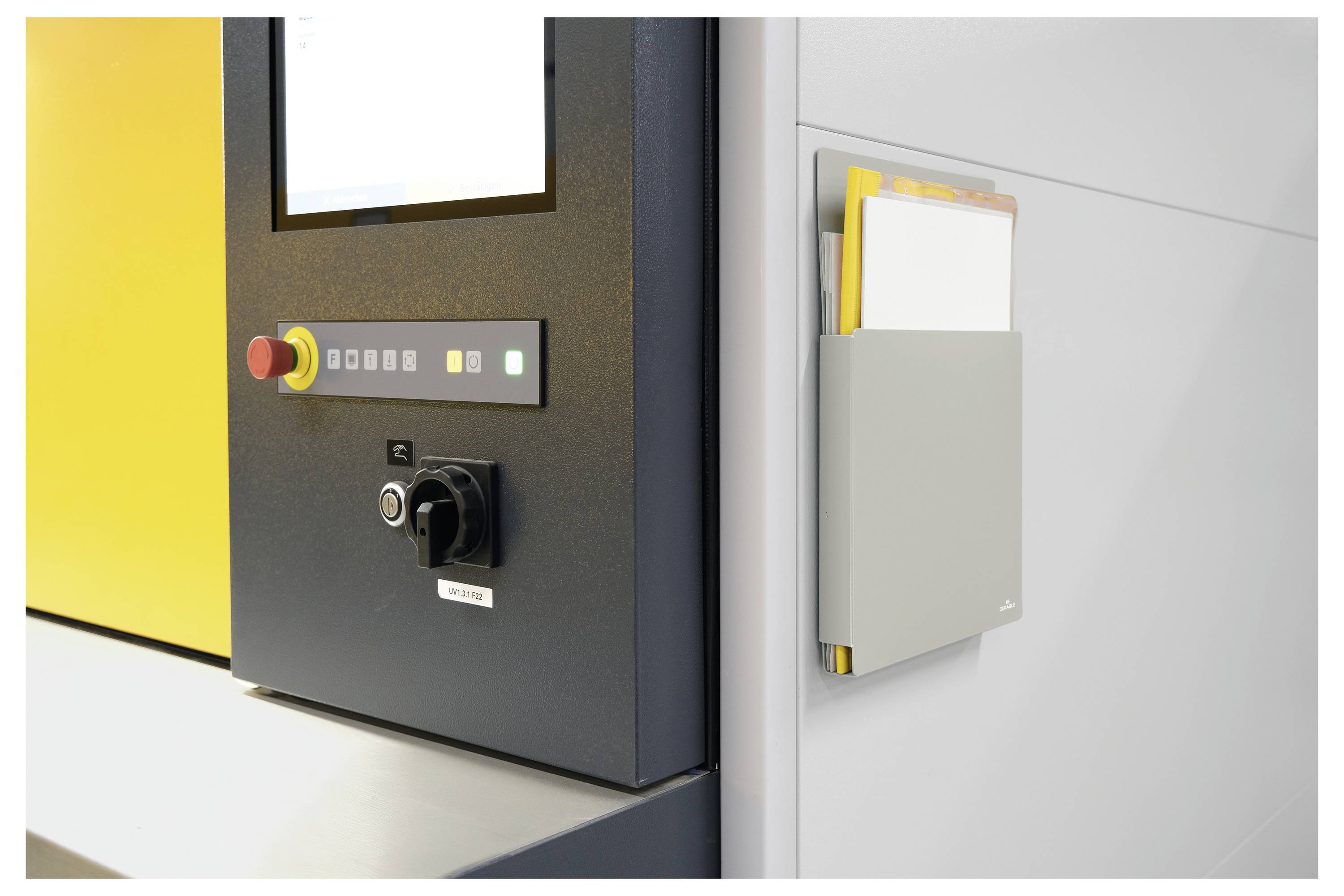 Nahaufnahme eines industriellen Bedienfeldes mit Notausstoppbutton und Schlüsselschalter neben einem an der Wand montierten Dokumentenhalter mit Papieren.