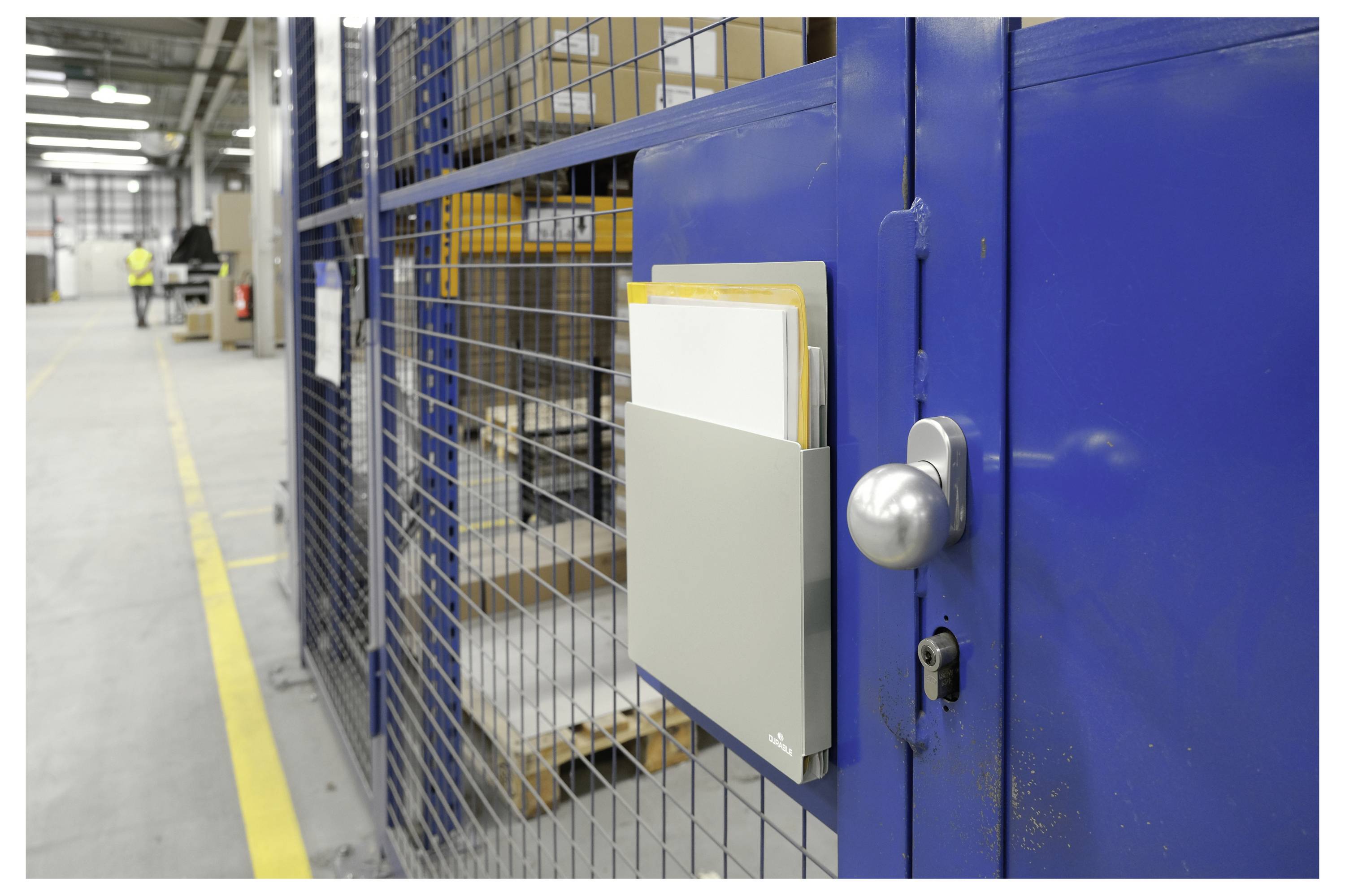 Eine blaue Sicherheitskäfigtür in einem Lagerhaus, ausgestattet mit einem Metallklemmbrett-Halter mit Dokumenten. Das Innere des Lagerhauses ist sichtbar.