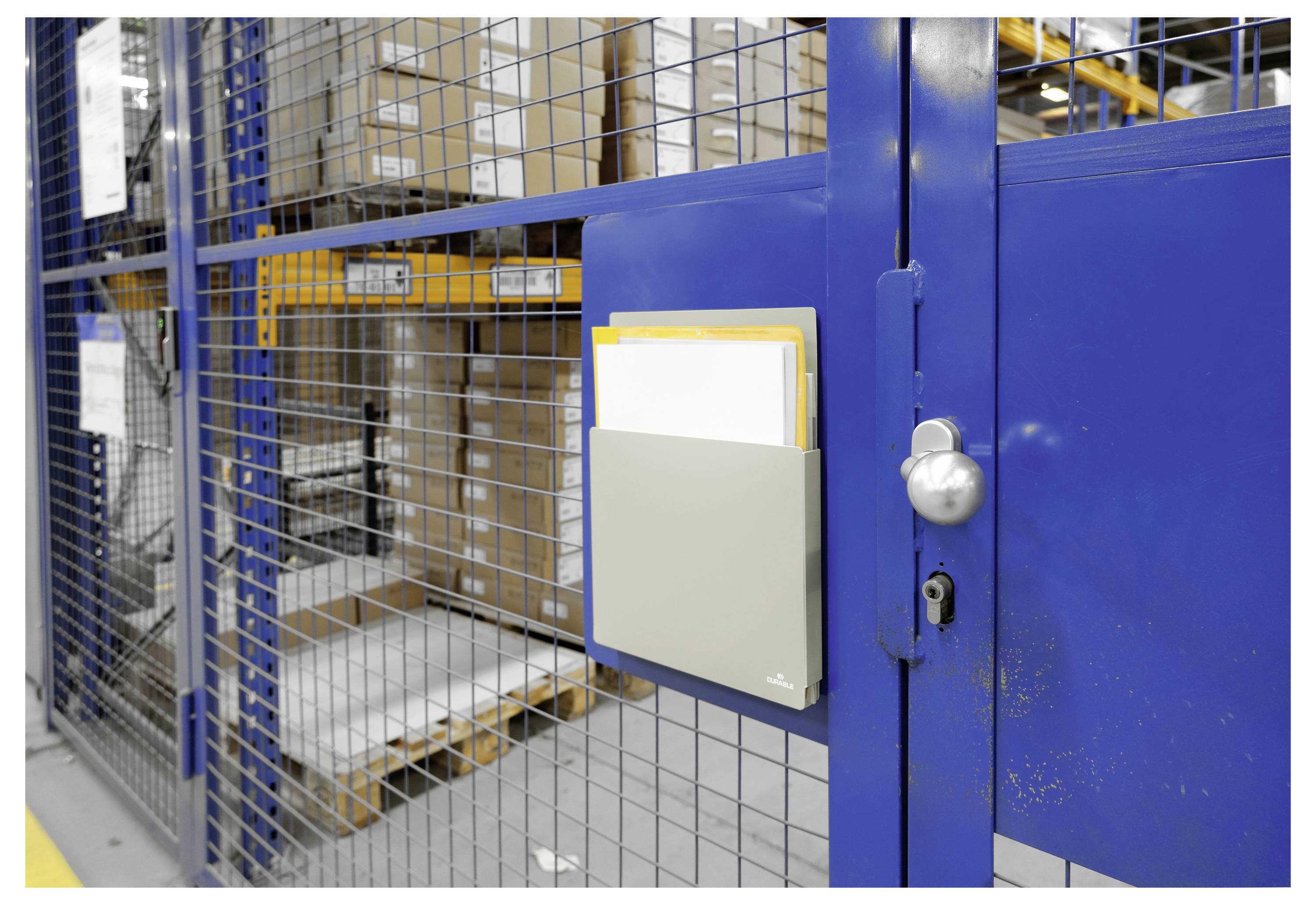 Dokumentenhalter an einem blauen Metalltor befestigt, mit Lagerkästen und Regalen im Hintergrund sichtbar durch einen Maschendrahtzaun.