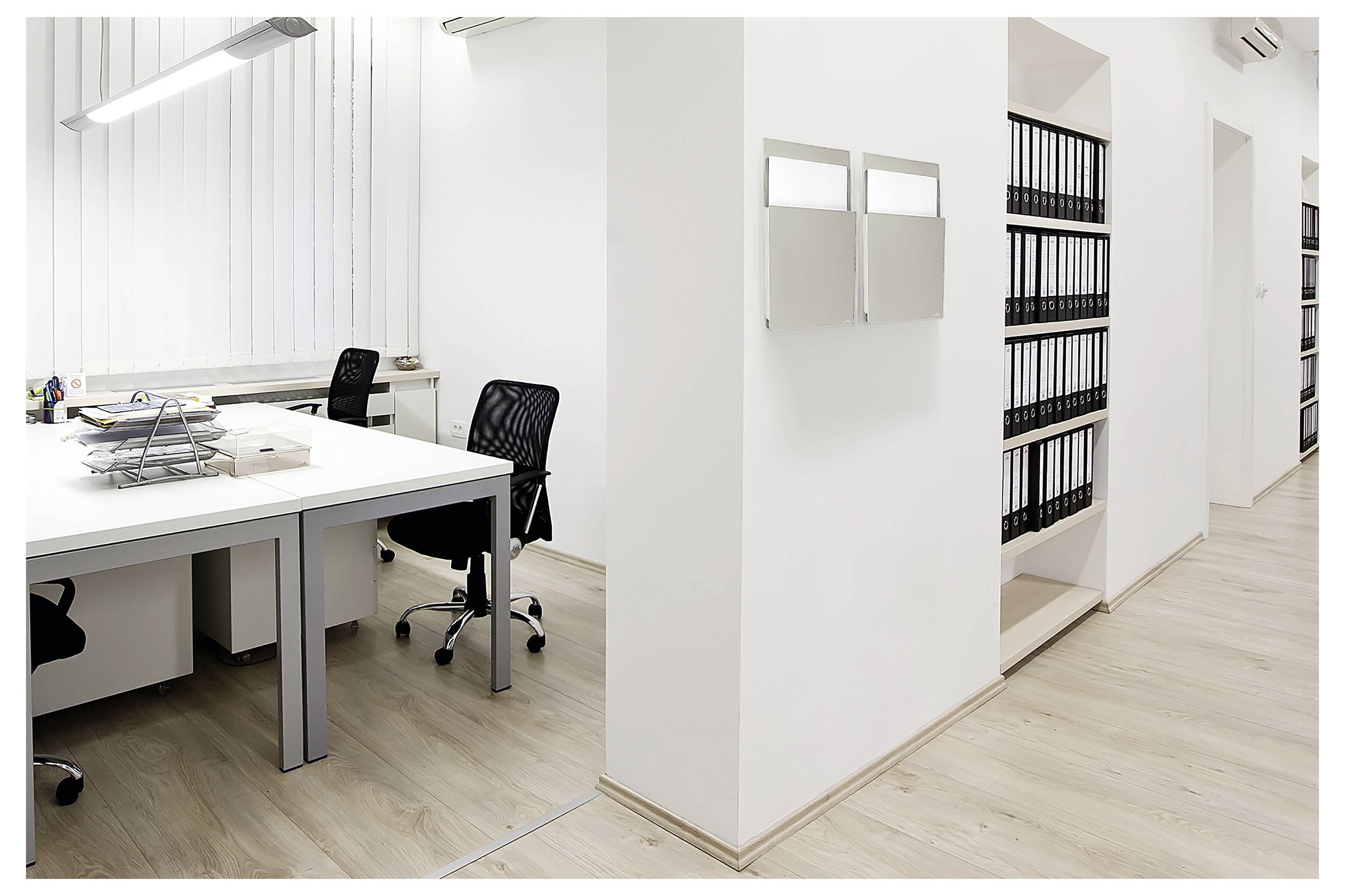 Modernes Büro mit zwei Schreibtischen und Stühlen, weißen Wänden, einem Stapel Papierwork auf einem Schreibtisch und Regalen gefüllt mit schwarzen Ordnern.