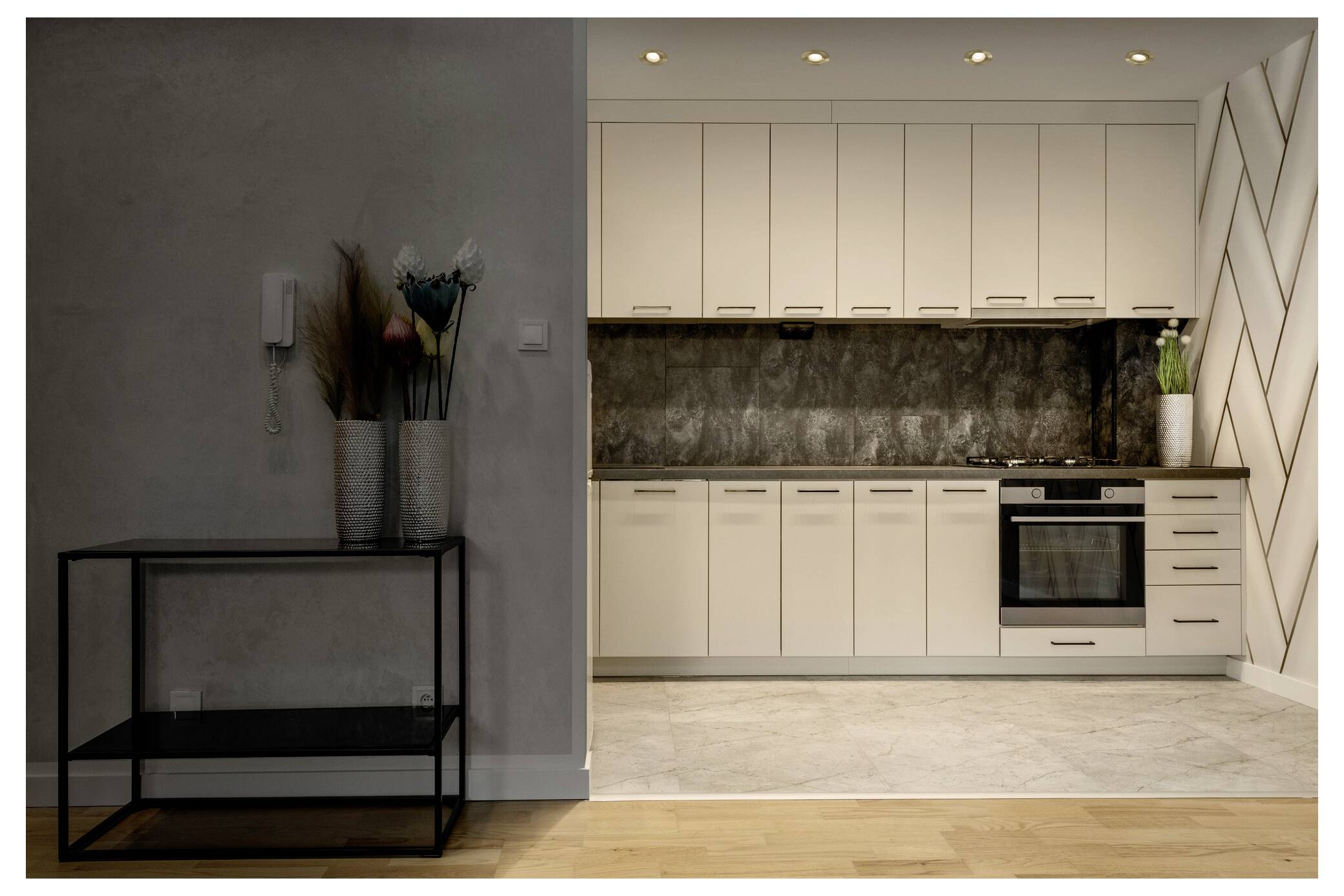 Eine moderne Küche mit weißen Schränken, einem Edelstahlofen und einer strukturierten Rückwand. Ein Regal mit Vasen und Blumen befindet sich auf der linken Seite.