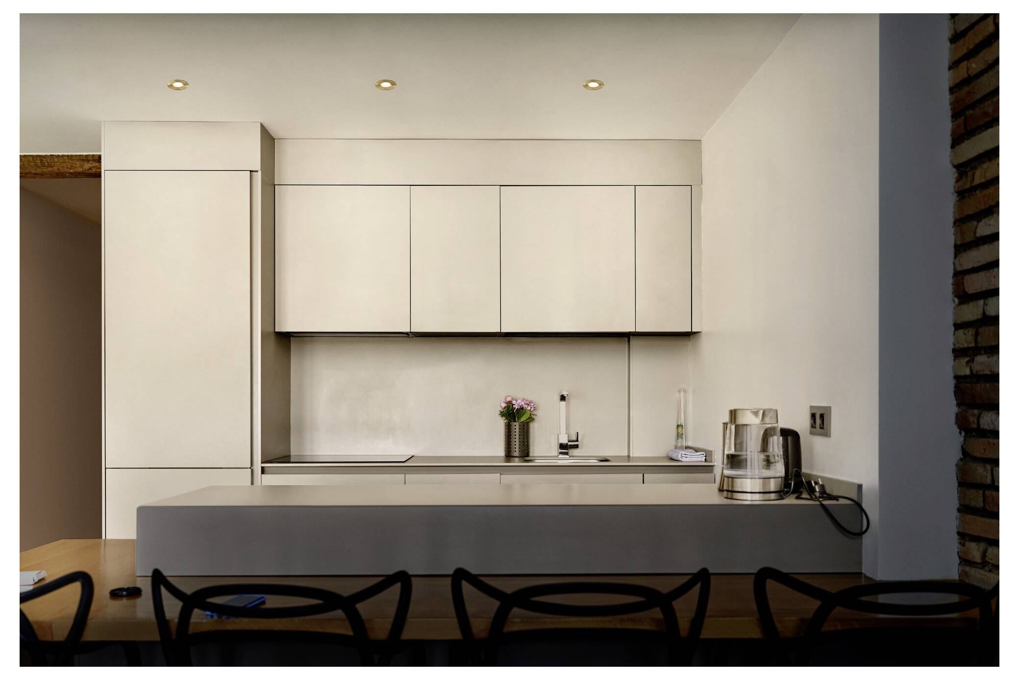 Moderne Küche mit heller Schrankwerk, eleganten Arbeitsplatten und einer Reihe schwarzer Stühle. Geräte und eine kleine Pflanze sind ordentlich angeordnet.