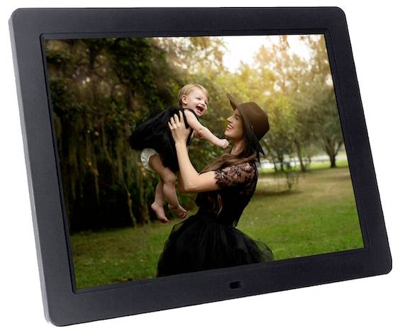 Eine Frau in einem schwarzen Kleid und Hut hebt freudig ein ähnlich gekleidetes Baby in einer Parklandschaft mit Bäumen und Gras hoch.