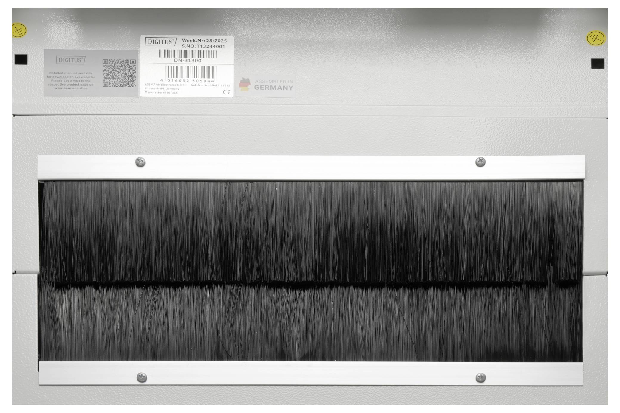 Das Bild zeigt ein Metallpanel mit einer bürstenartigen Öffnung, wahrscheinlich Teil einer Industriemaschine. Beschriftungen und ein QR-Code sind auf dem Panel sichtbar.